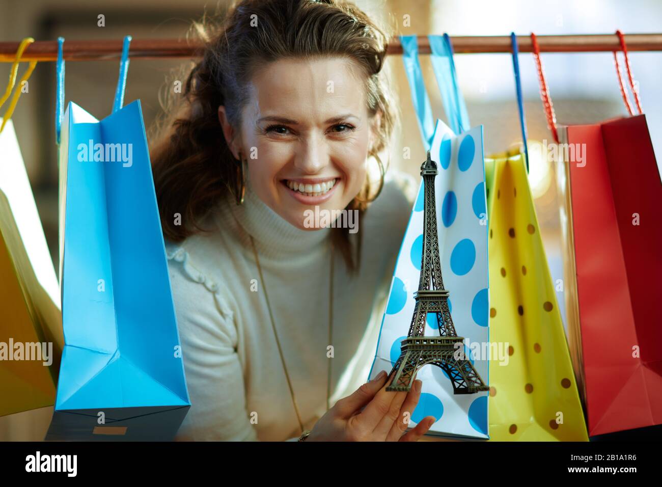 Jeune femme souriante dans un pull blanc et une jupe à emporter avec des sacs de shopping colorés accrochés sur un rail en cuivre avec souvenir de la tour Eiffel. Banque D'Images