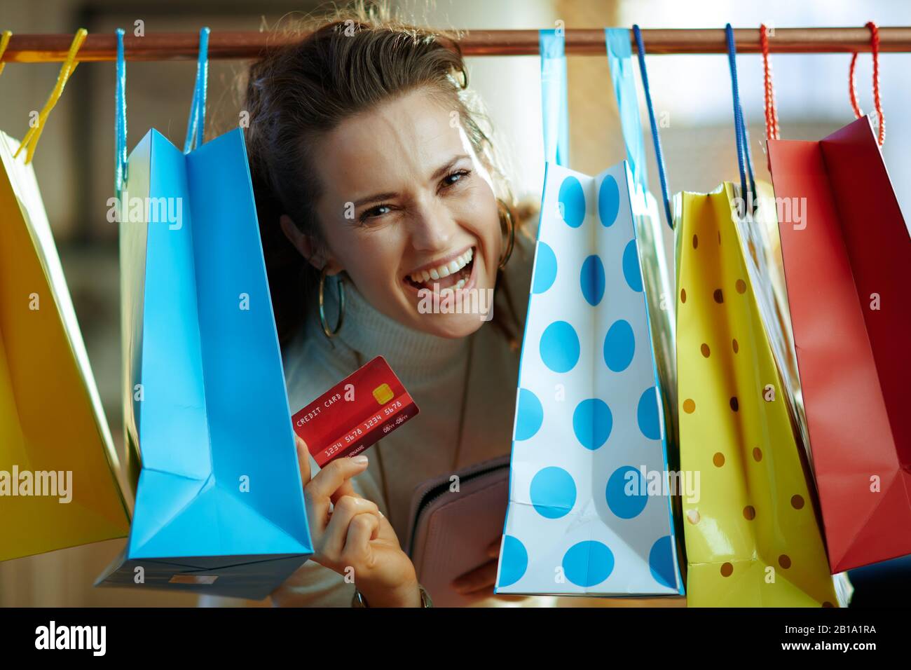 jeune femme souriante dans un pull blanc et une jupe qui s'épluche des sacs à provisions colorés accrochés sur un rail en cuivre avec carte de crédit rouge et sac à main. Banque D'Images