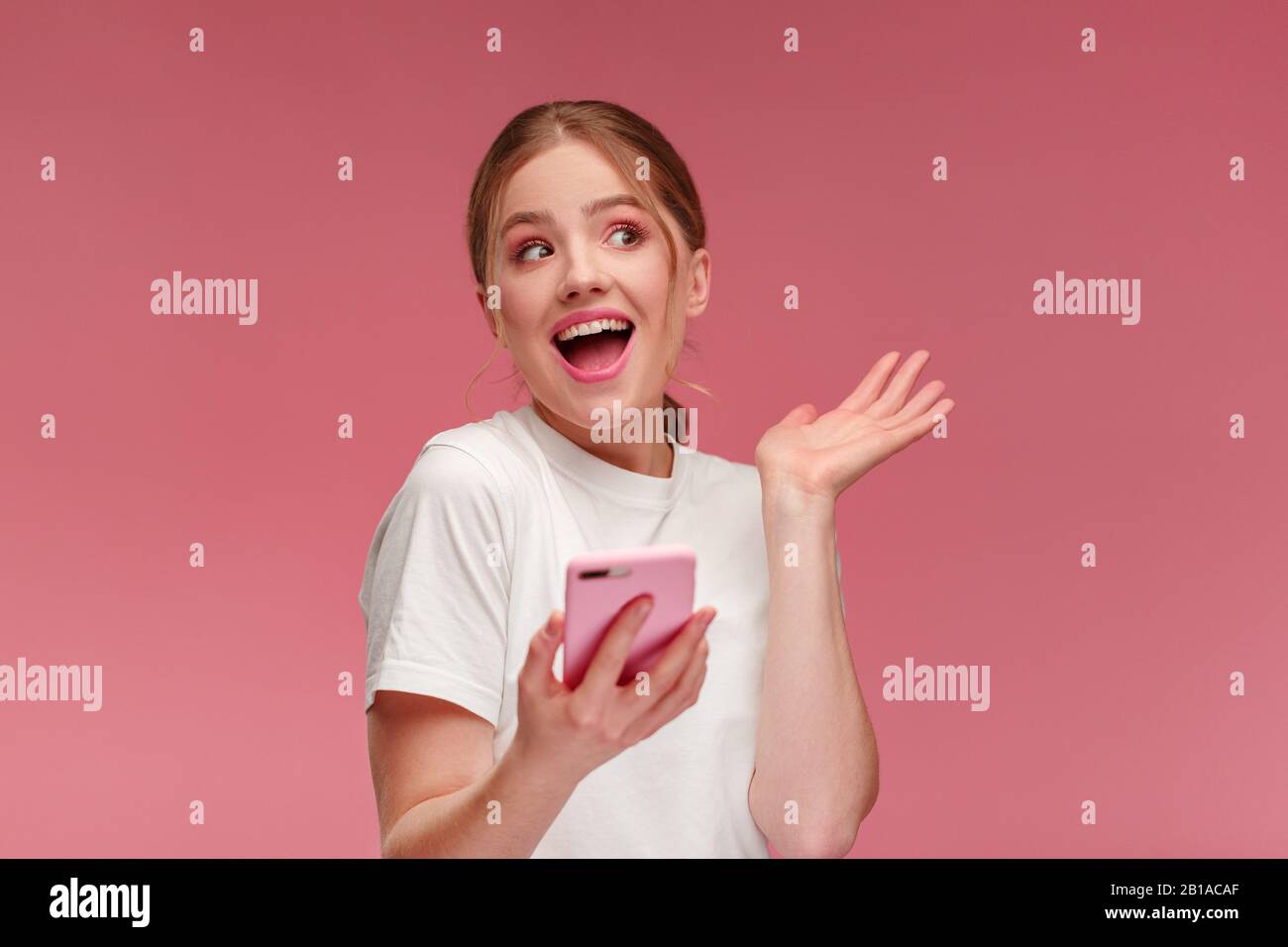 La meilleure journée de notre vie. Jeune femme à tête rouge surparie tenant un smartphone rose, souriant et exprimant sa positivité. Bonne fille a reçu des nouvelles positives choquantes. Cop Banque D'Images