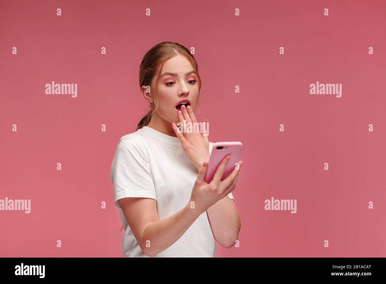 La meilleure journée de notre vie. Jeune femme à tête rouge surparie tenant un smartphone rose, souriant et exprimant sa positivité. Bonne fille a reçu des nouvelles positives choquantes. Cop Banque D'Images