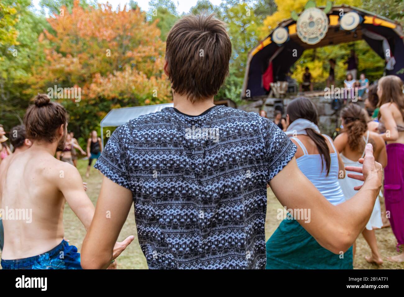 Une vingtaine de boho gars est vu de l'arrière, avec des cheveux bruns et un t-shirt imprimé bleu, dansant au festival avec des personnes floues en arrière-plan Banque D'Images