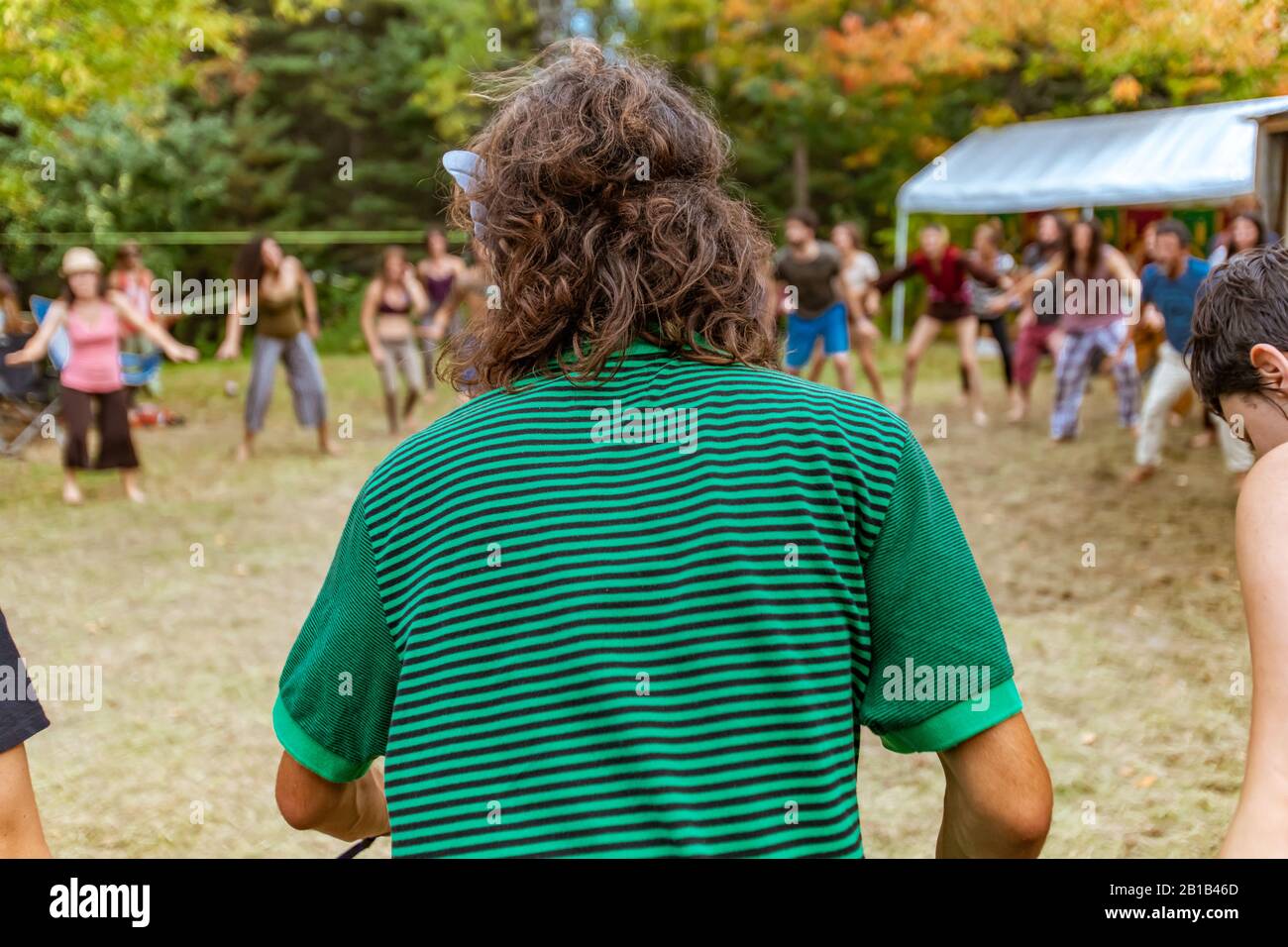 Un homme bohème avec des cheveux de brunette ondulés de longueur d'épaule est vu de derrière dans un point peu profond, portant un t-shirt à rayures vertes au festival multiculturel Banque D'Images