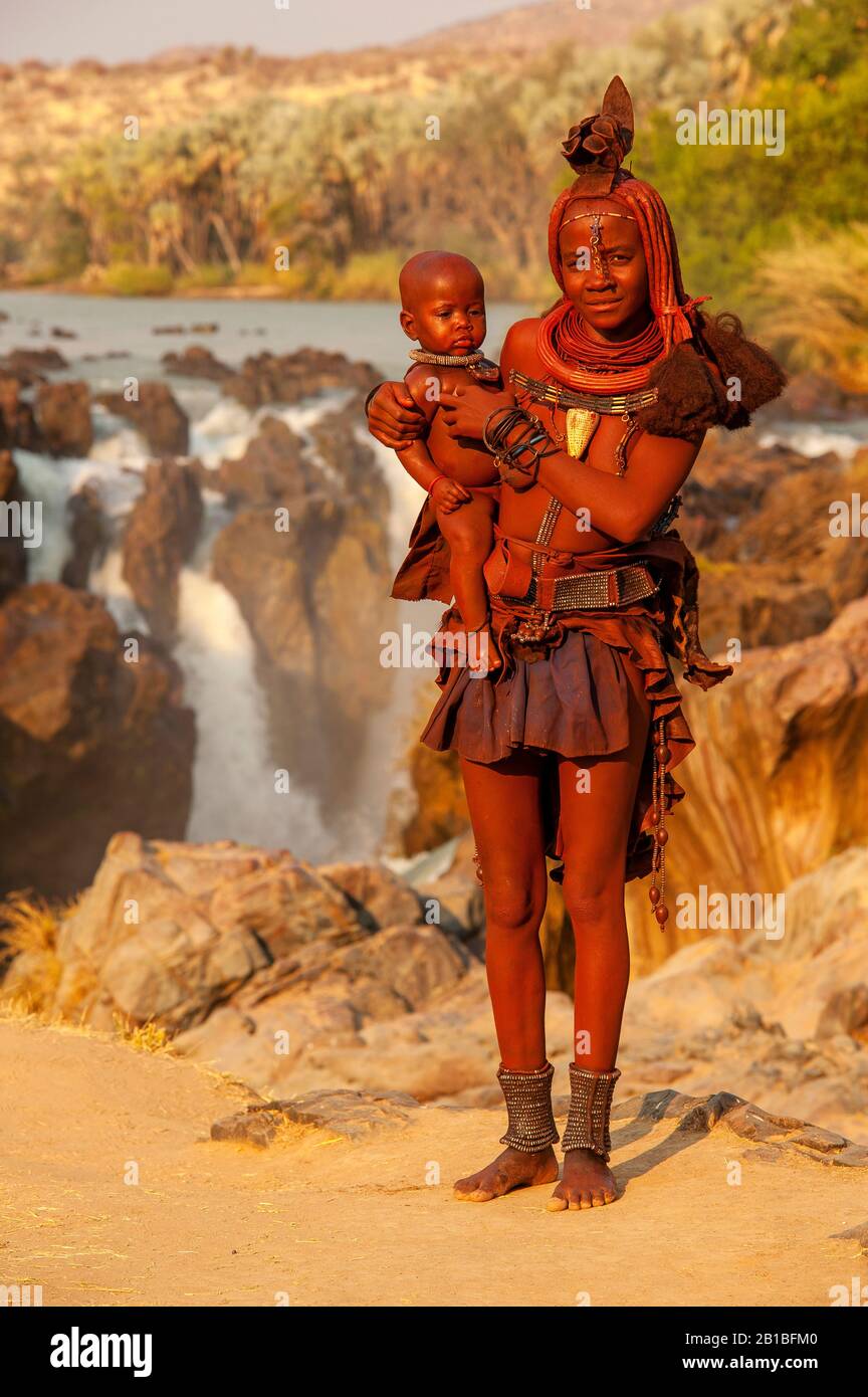 Himba femme sport son cou plein d'ornements traditionnels et un petit bébé sur ses genoux, Epupa Falls en arrière-plan, région de Kunene, Namibie Banque D'Images