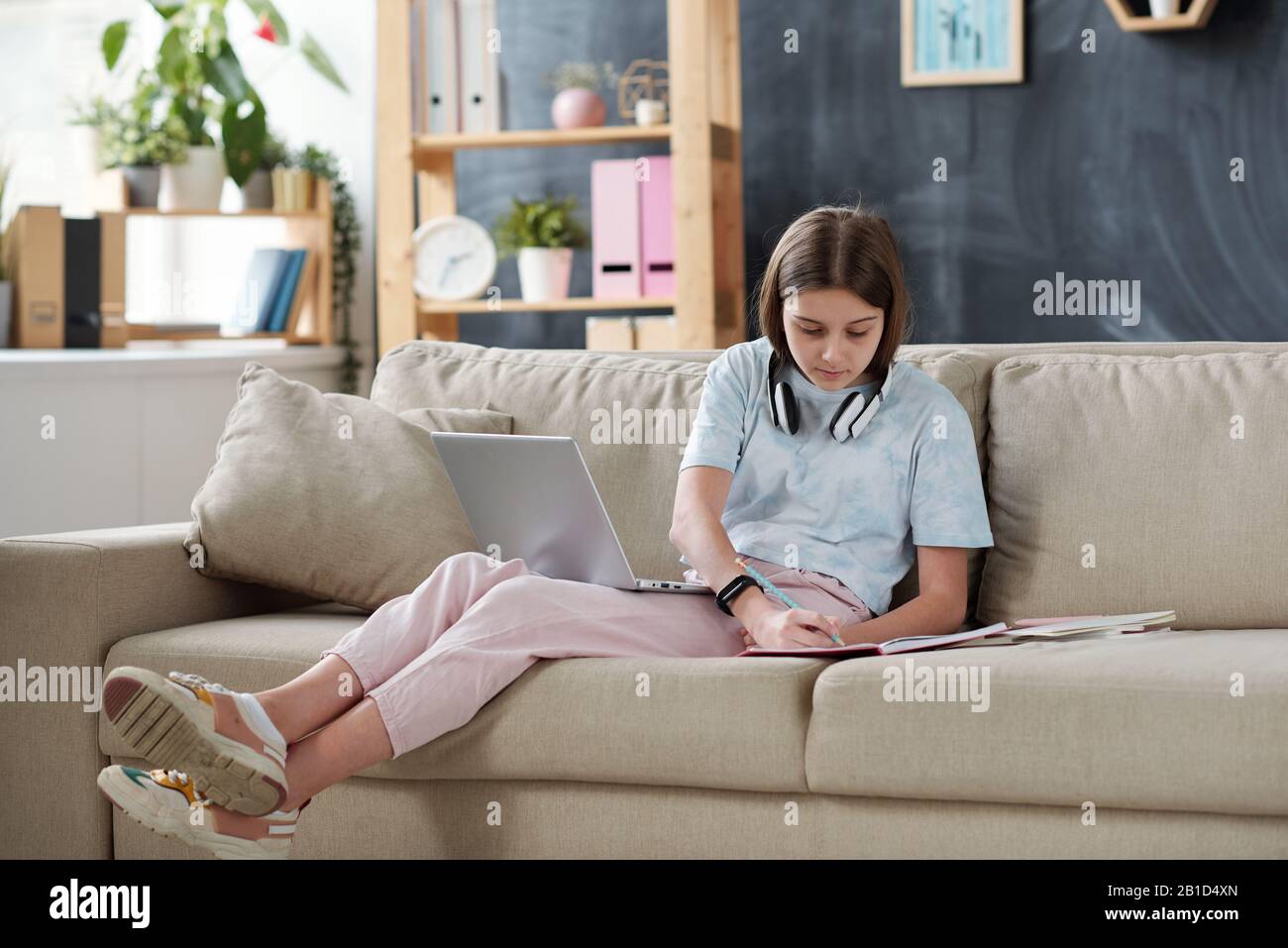 Une adolescente avec écouteurs sur le cou utilisant des ressources Internet sur un ordinateur portable tout en faisant des devoirs dans le salon Banque D'Images