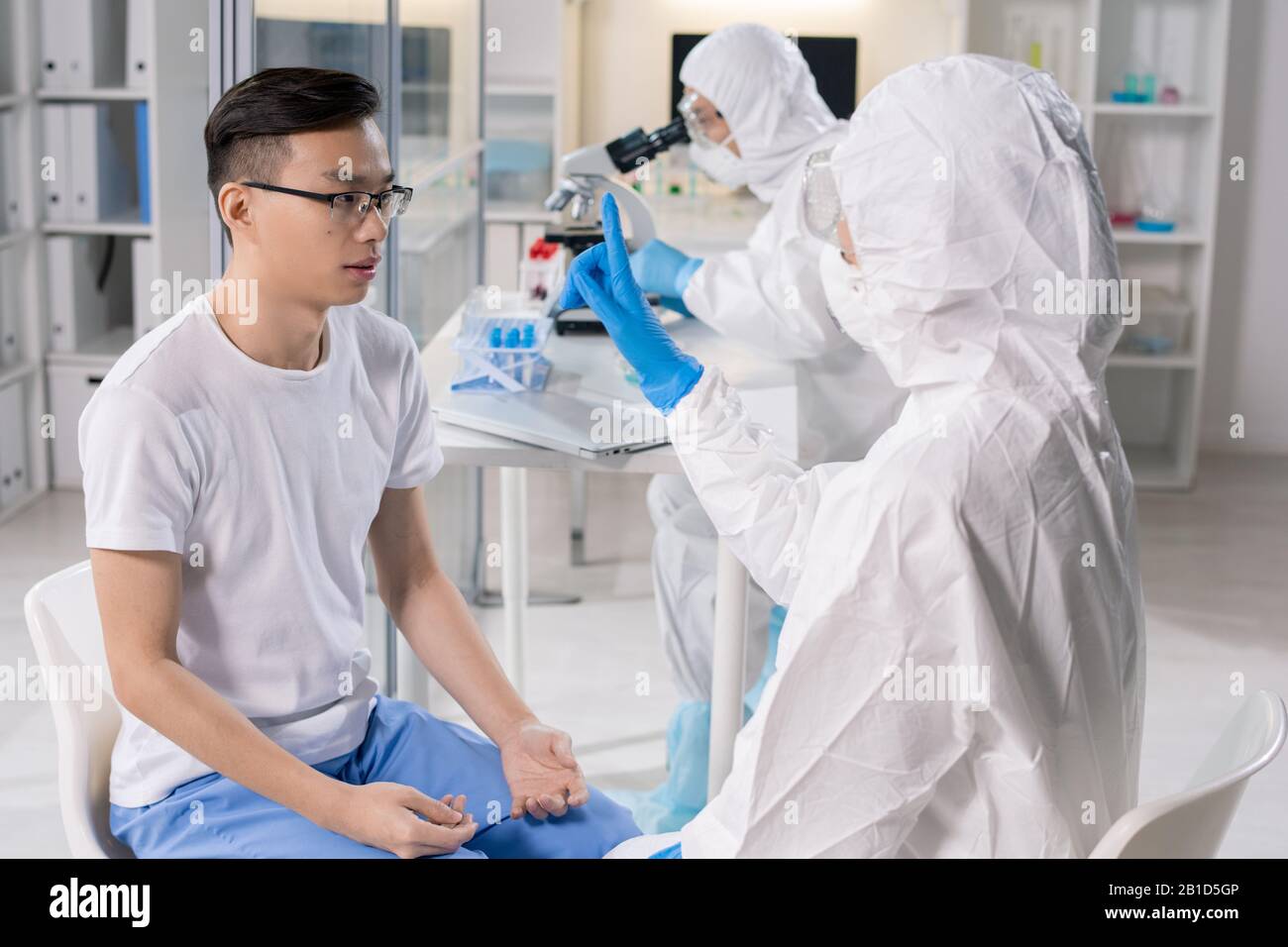 Spécialiste médical en costume protecteur montrant le doigt au patient asiatique infecté tout en vérifiant sa réaction Banque D'Images