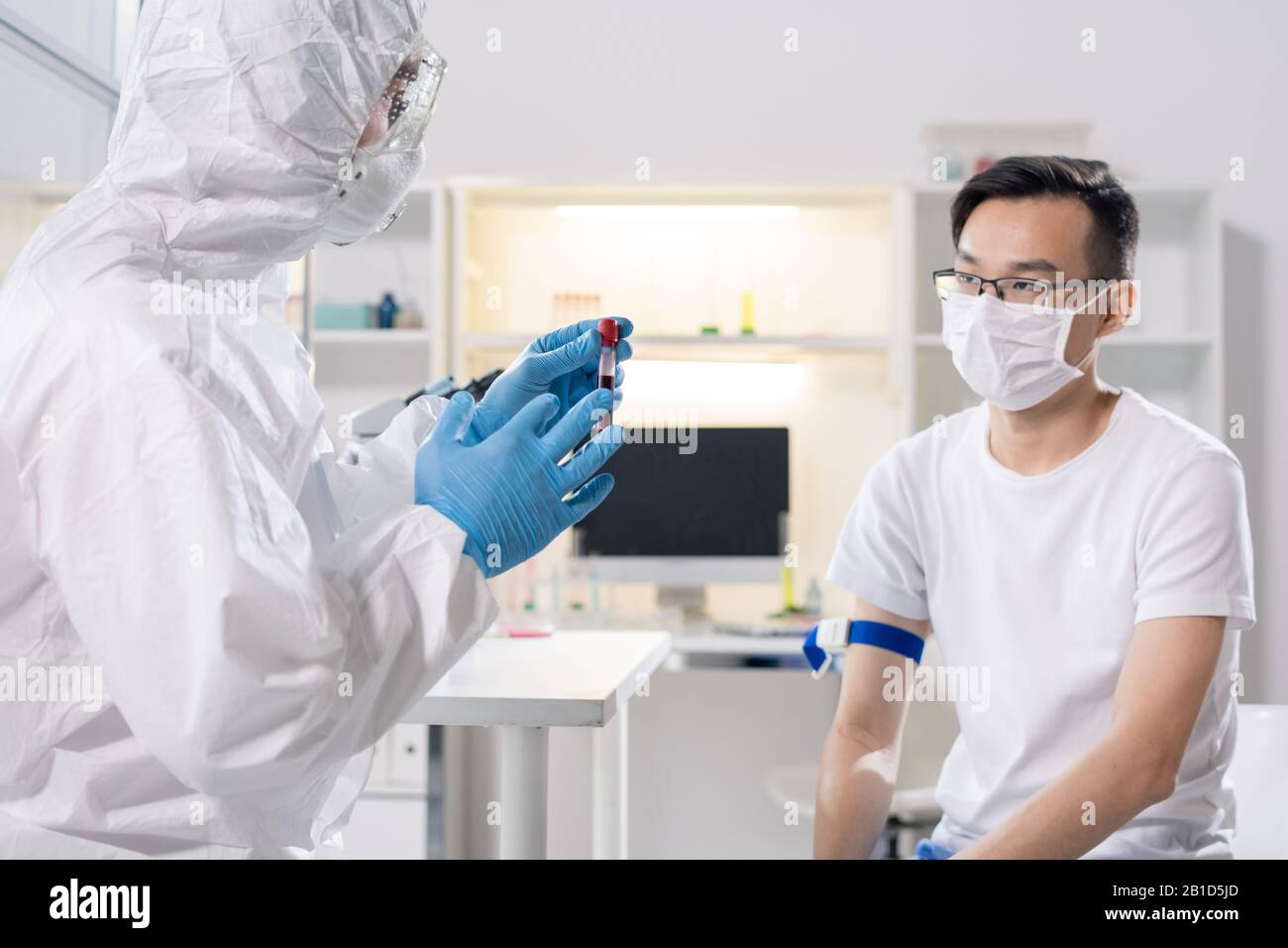 Spécialiste médical en costume de protection parlant au patient asiatique tout en prenant du sang pour la recherche en laboratoire Banque D'Images