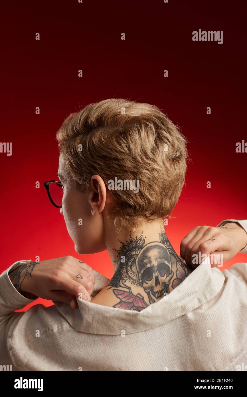 Vue arrière de la jeune femme à poil court avec tatouage au cou avec crâne et papillons posant sur fond rouge en studio Banque D'Images