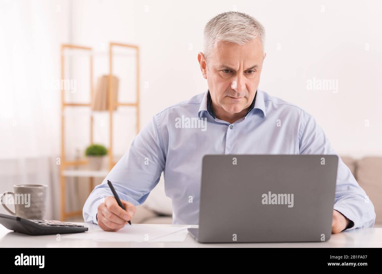 Homme senior utilisant un ordinateur portable au bureau et écrivant des notes Banque D'Images