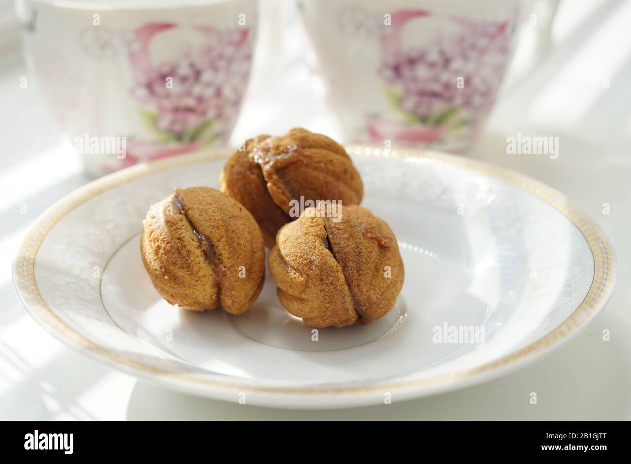 Trois petits cookies sous forme de noix sur une soucoupe blanche. Arrière-plan deux tasses de thé sur une table ensoleillée. Banque D'Images