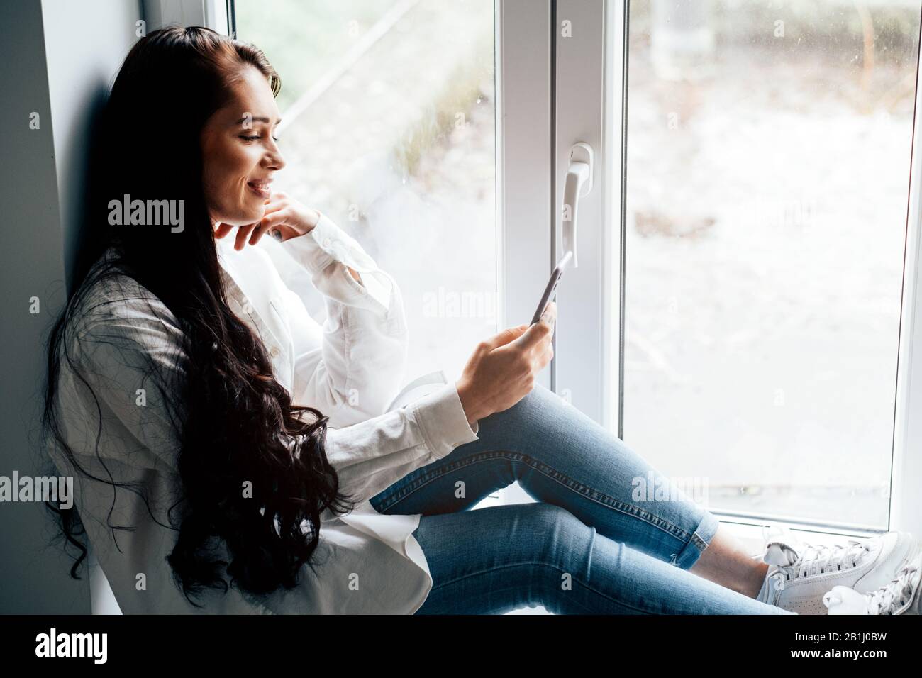jolie jeune femme souriante assise sur le seuil de la fenêtre à l'aide d'un téléphone mobile Banque D'Images
