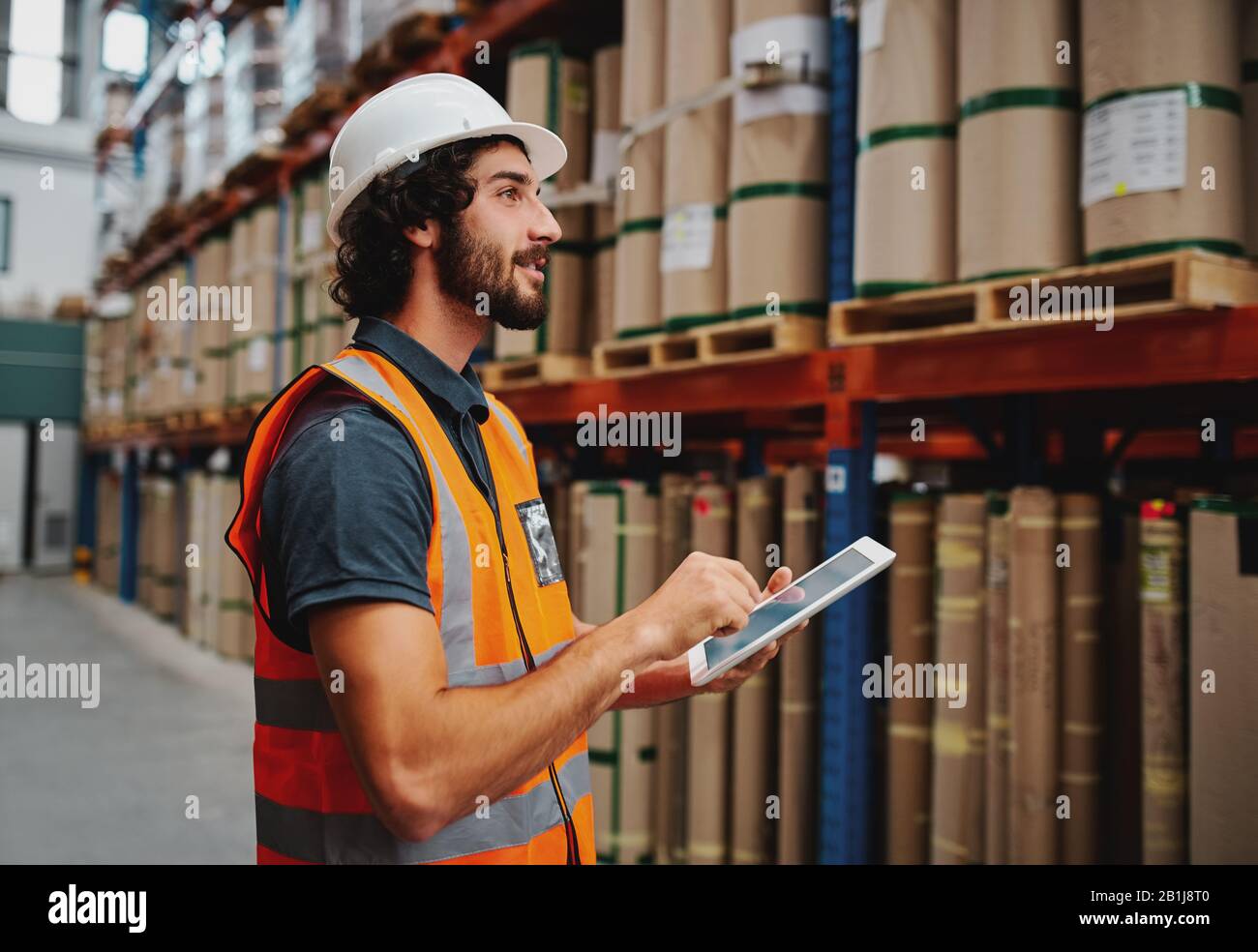 Jeune employé en entrepôt dans un casque et une veste de sécurité utilisant une tablette numérique tout en prenant commande et en confirmant la disponibilité des stocks à l'aide d'une tablette numérique Banque D'Images