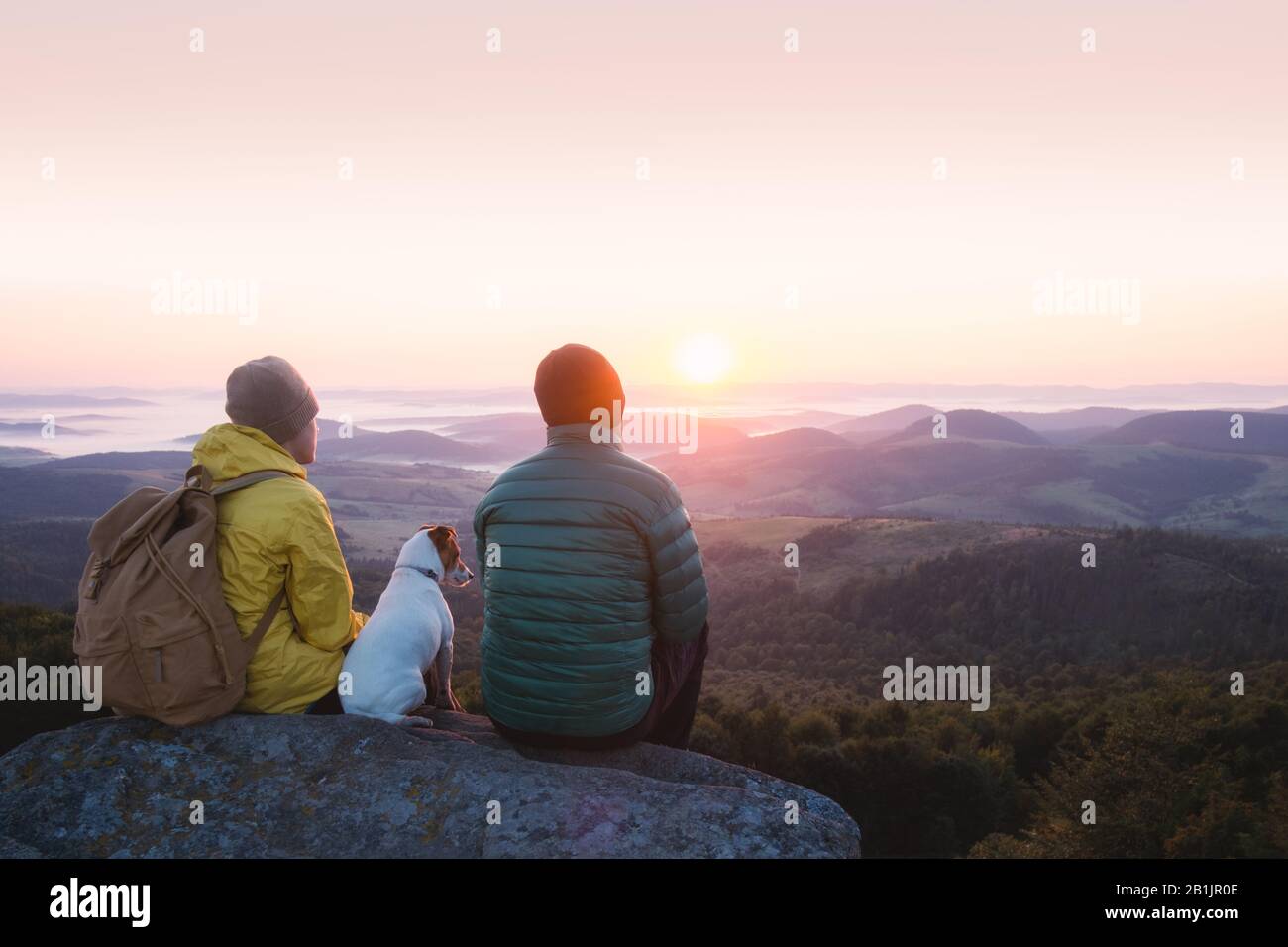 Deux touristes assis sur le bord de la falaise avec chien blanc sur fond d'une incroyable montagne de lever de soleil. Photographie de paysage Banque D'Images