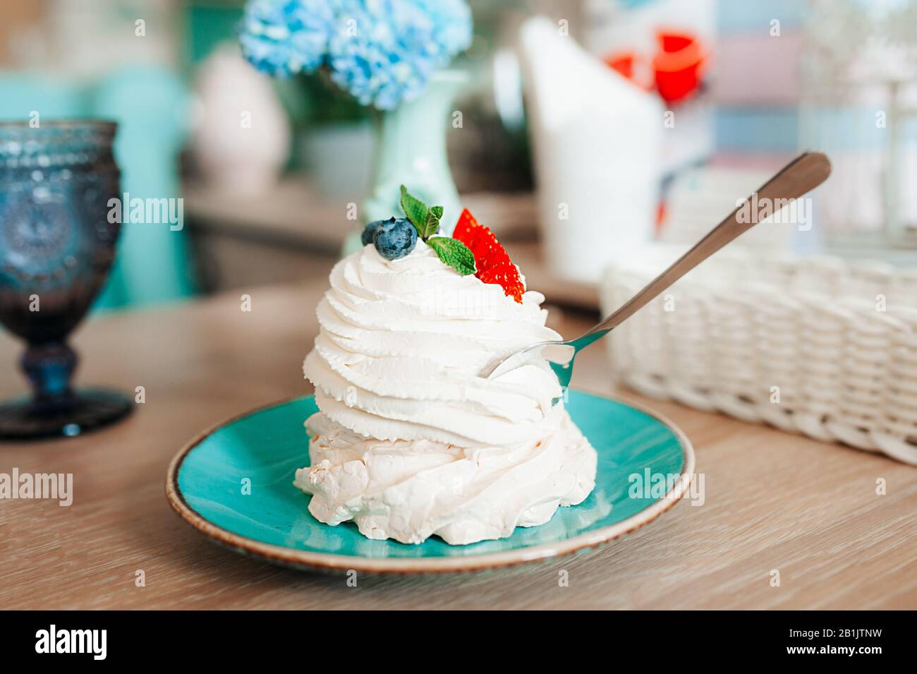 Dessert blanc et spacieux de meringue et crème avec fraises et bleuets sur une plaque bleue à l'intérieur d'un café lumineux. La cuillère argentée tente Pavlov Banque D'Images