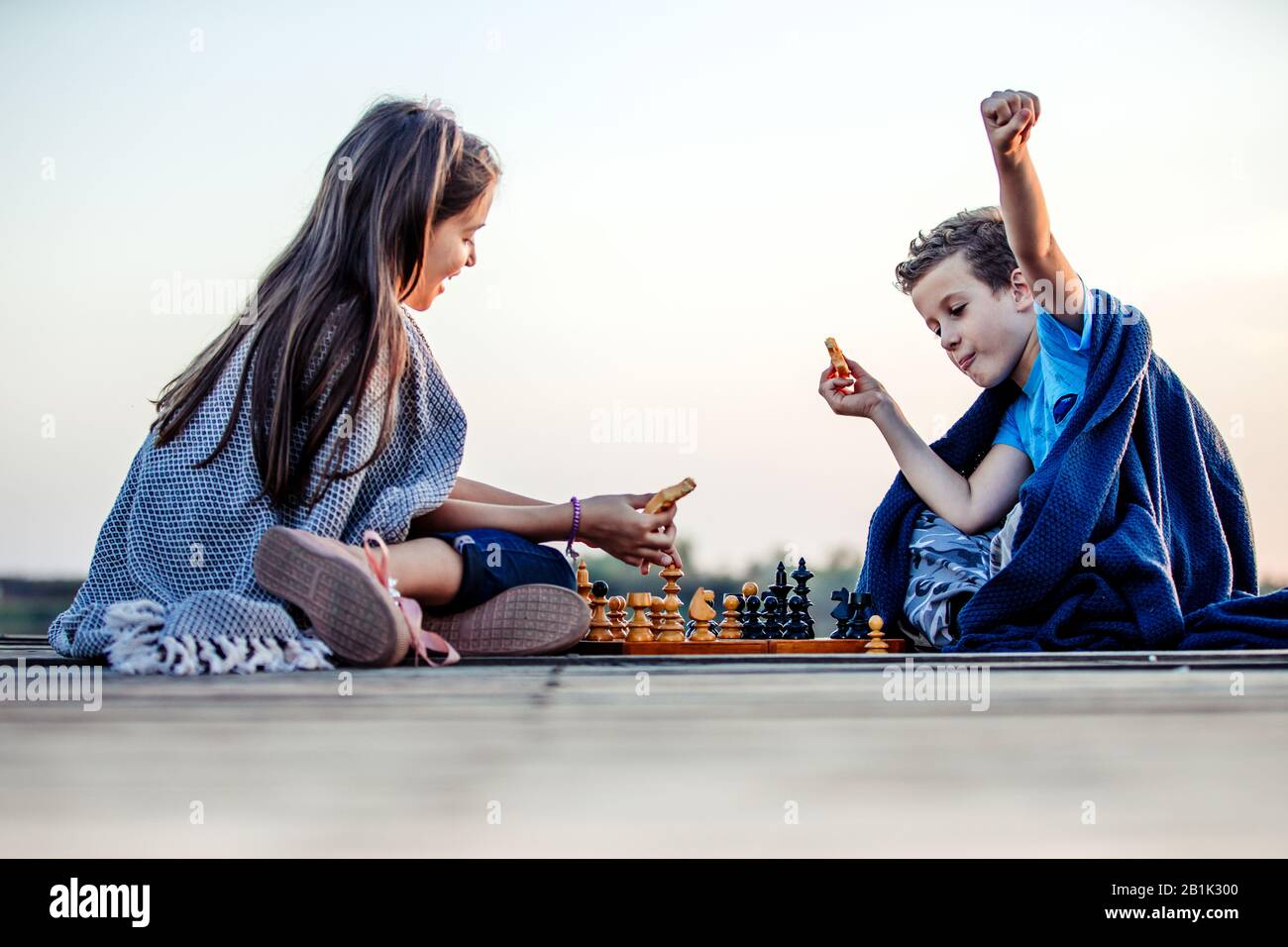 Deux jeunes petits amis mignons, un garçon et une fille qui s'amusent en jouant aux échecs assis près du lac le soir. Les enfants jouent.geste gagnant. Ami Banque D'Images