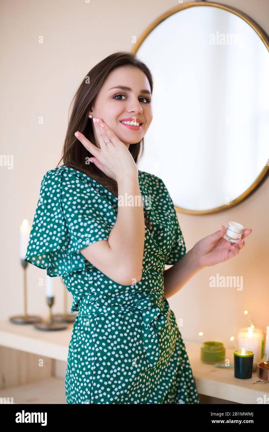 Brunette heureuse dans la polka verte robe à pois hydratant la peau du visage avec crème regardant l'appareil photo Banque D'Images