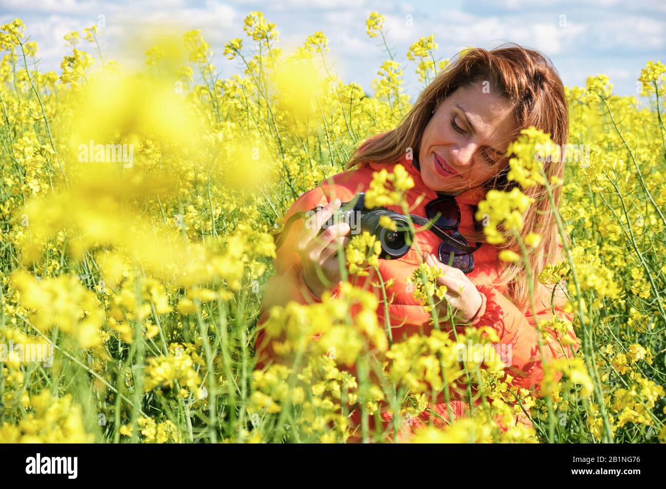 Belle femme photographe souriant et regardant son appareil photo, à l'intérieur d'un champ de colza jaune. Banque D'Images