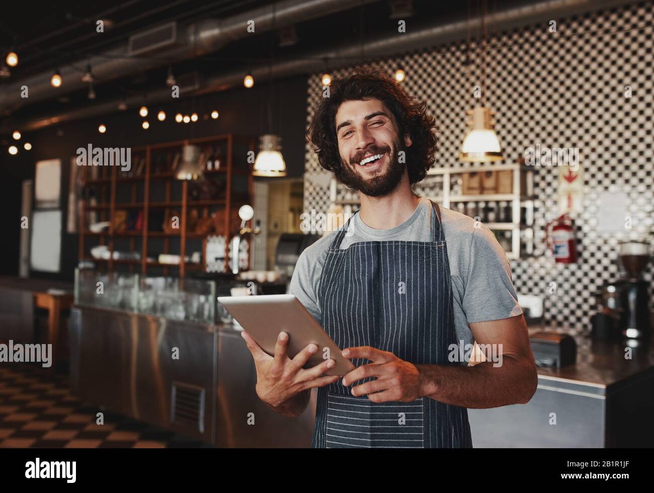 Beau jeune propriétaire de café caucasien portant un tablier riant à l'aide d'une tablette numérique Banque D'Images