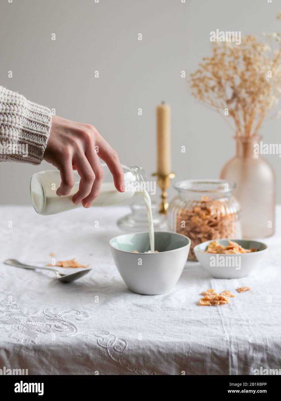 La main de la femme verse du lait dans un bol avec des cornflakes. Table lumineuse du matin avec fleurs séchées Banque D'Images