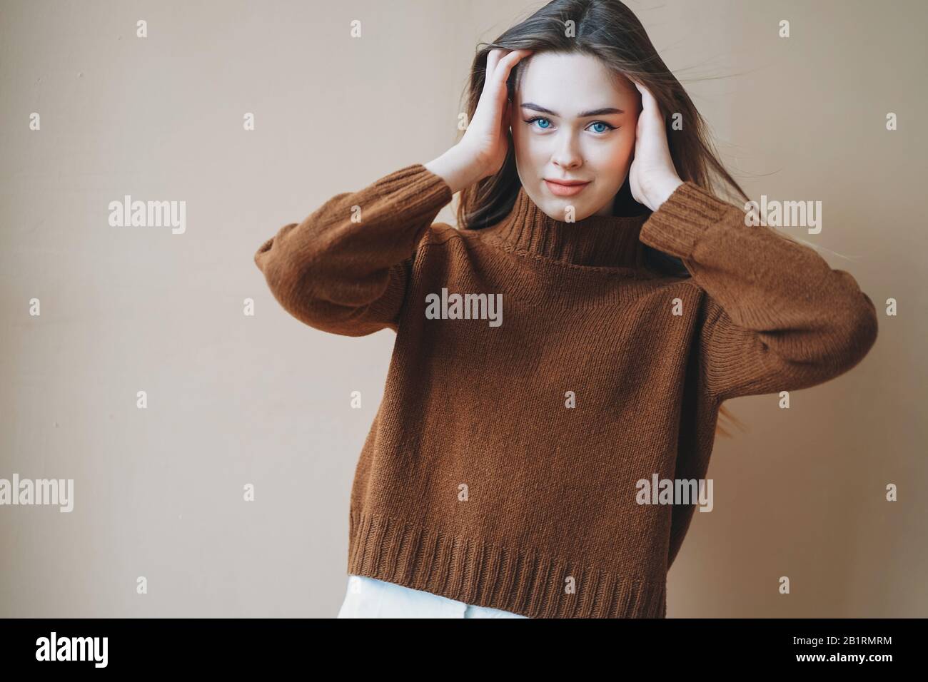 Jeune fille de cheveux longue à poil brun avec yeux bleus dans un pull tricoté brun regardant l'appareil photo sur le fond beige Banque D'Images