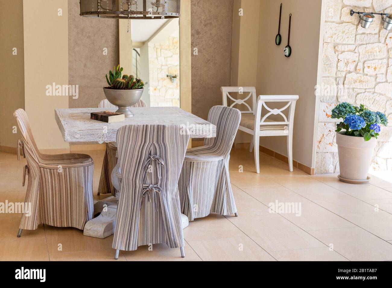 Chaises avec couvertures en tissu rayé autour de la table carrée, banc blanc, hortensia bleu dans pot à fleurs. Intérieur de salon de style méditerranéen Banque D'Images