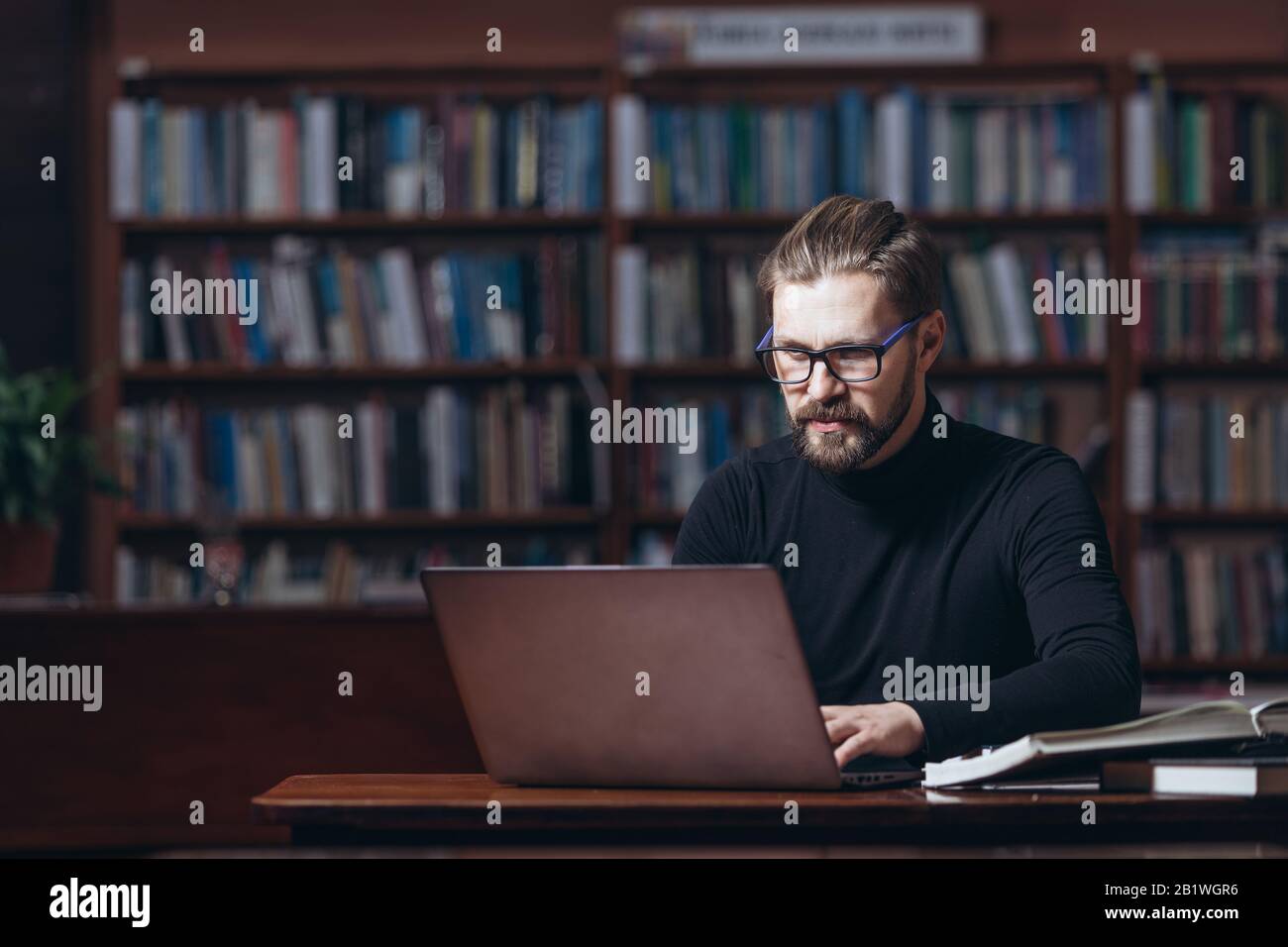 Homme sérieux barbu à col polo noir assis au bureau dans la bibliothèque publique et travaillant sur un ordinateur portable personnel. Homme confiant dans les lunettes qui travaillent sur la prise de vue Banque D'Images