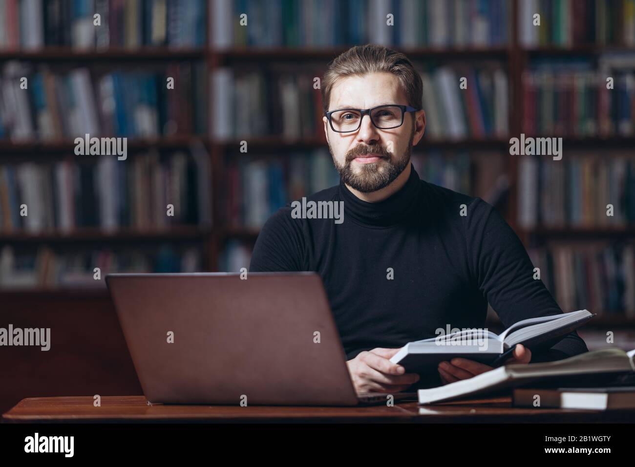 Homme confiant dans le col polo noir assis à la table avec livre ouvert et ordinateur portable et regardant l'appareil photo. Homme mûr dans des lunettes travaillant au public Banque D'Images