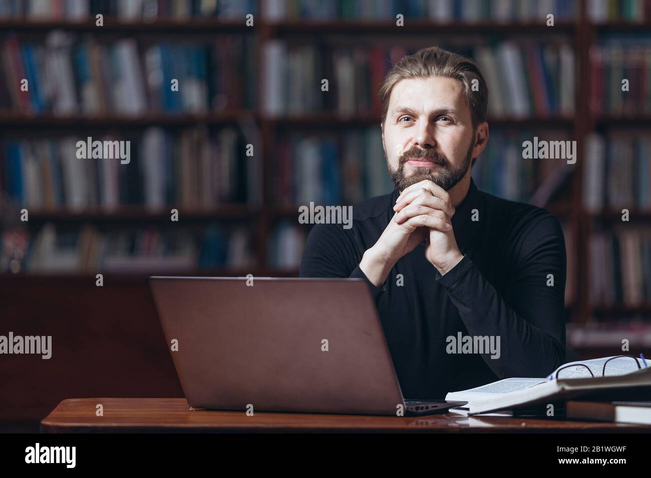 Homme mûr barbu à col polo noir assis à la table avec des livres ouverts et regardant l'appareil photo avec fond flou. Un homme heureux a fini de travailler sur le revers Banque D'Images