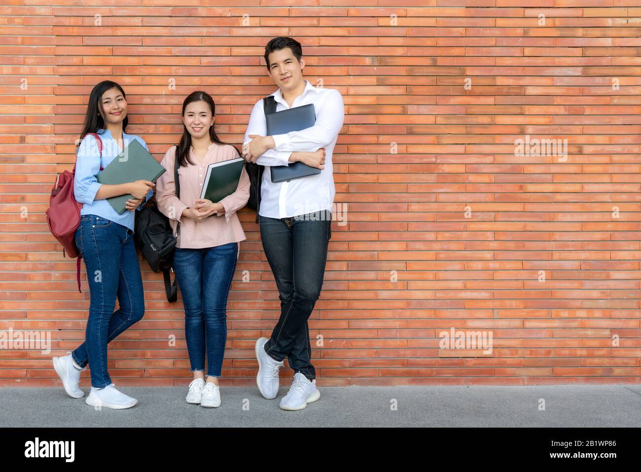Trois étudiants asiatiques souriants tenant livre et ordinateur portable posant sur fond de brique dans le campus. Groupe de jeunes élèves de l'école secondaire à l'extérieur. Banque D'Images