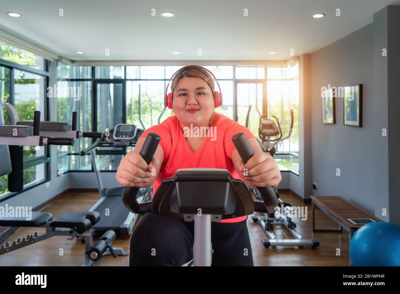 Heureuse femme asiatique En Surpoids avec une formation d'écouteurs sur vélo d'exercice dans la salle de sport moderne, heureux et sourire pendant l'entraînement. Femme grasse prendre soin de la santé et Banque D'Images