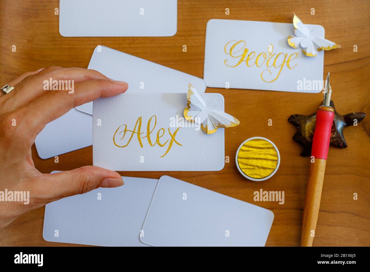 Calligraphie de mariage - main féminine tenant des cartes de lieu manuscrites avec des noms et des papillons en papier. Écriture dorée sur papier blanc. Banque D'Images