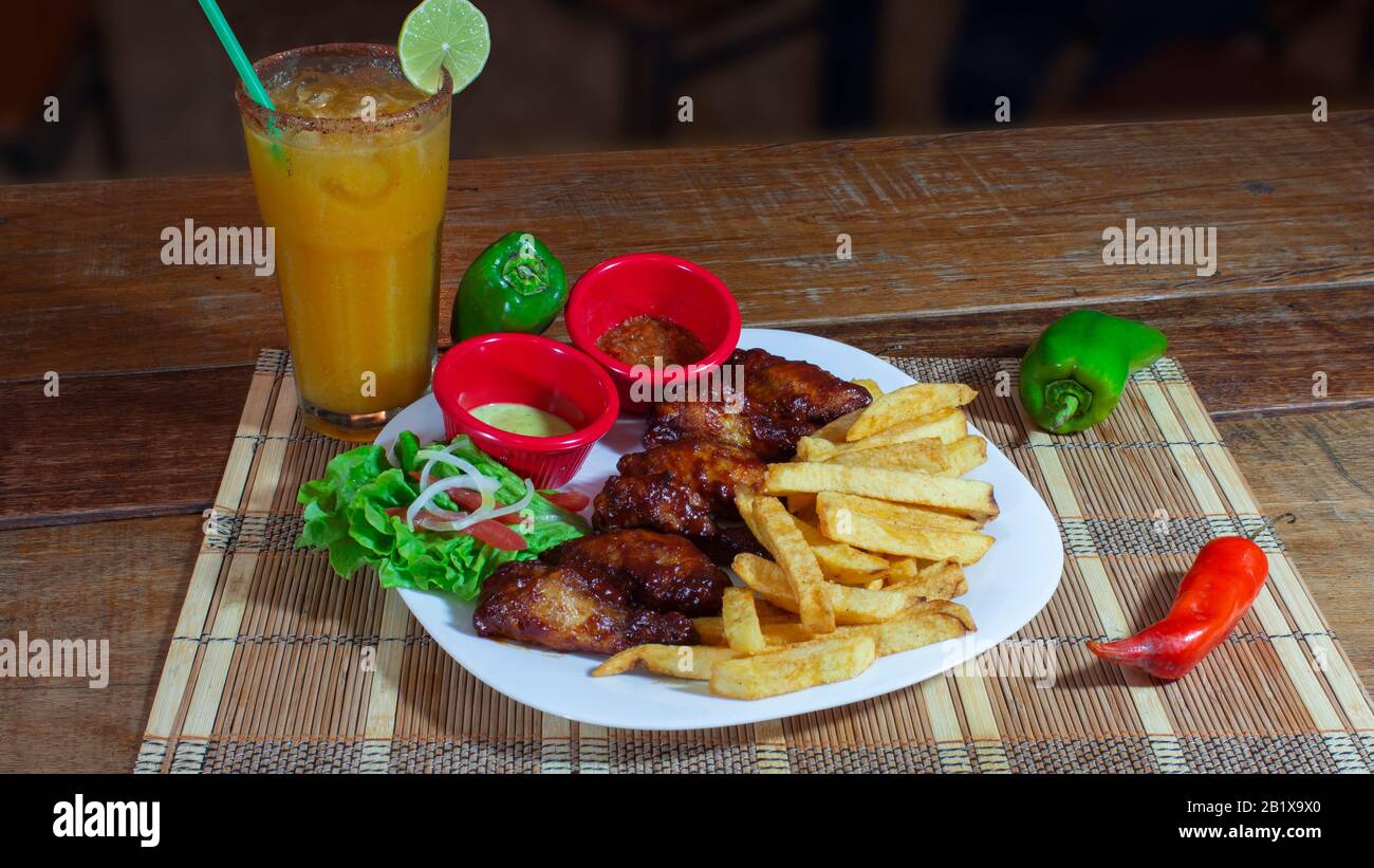 Plaque blanche avec ailes de poulet barbecue accompagnées de frites, sauces et salade sur une table en bois garnie de poivrons verts et rouges et d'un verre de Banque D'Images
