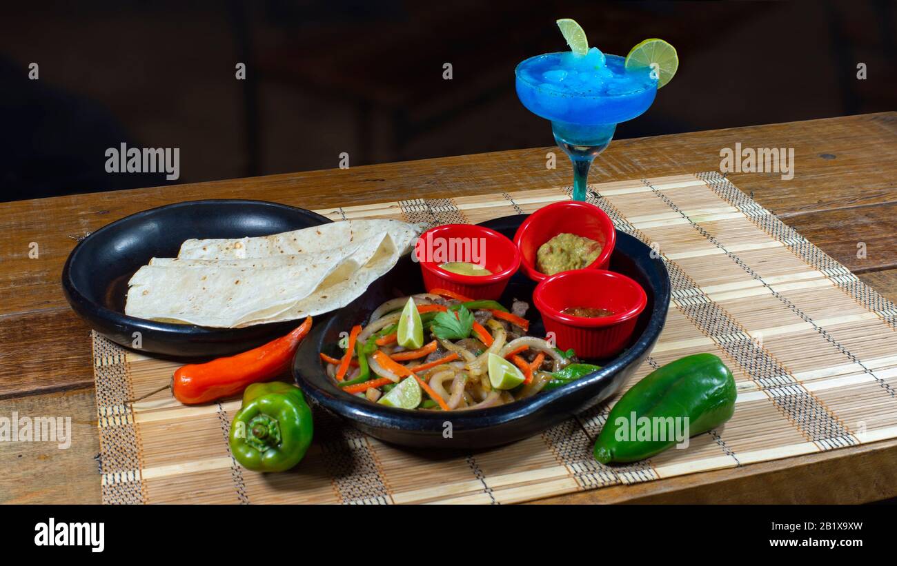 Plat d'argile avec fajitas de boeuf avec des morceaux de poivre et d'oignon accompagnés d'une plaque de tortillas de maïs et de sauce épicée, guacamole et une marguerite bleue sur un Banque D'Images