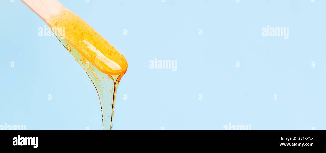 la cire jaune liquide ou la pâte de sucre pour l'épilation s'écoule du bâton sur fond bleu. Le concept d'épilation, de cirage, de sugaring peau lisse avec Banque D'Images