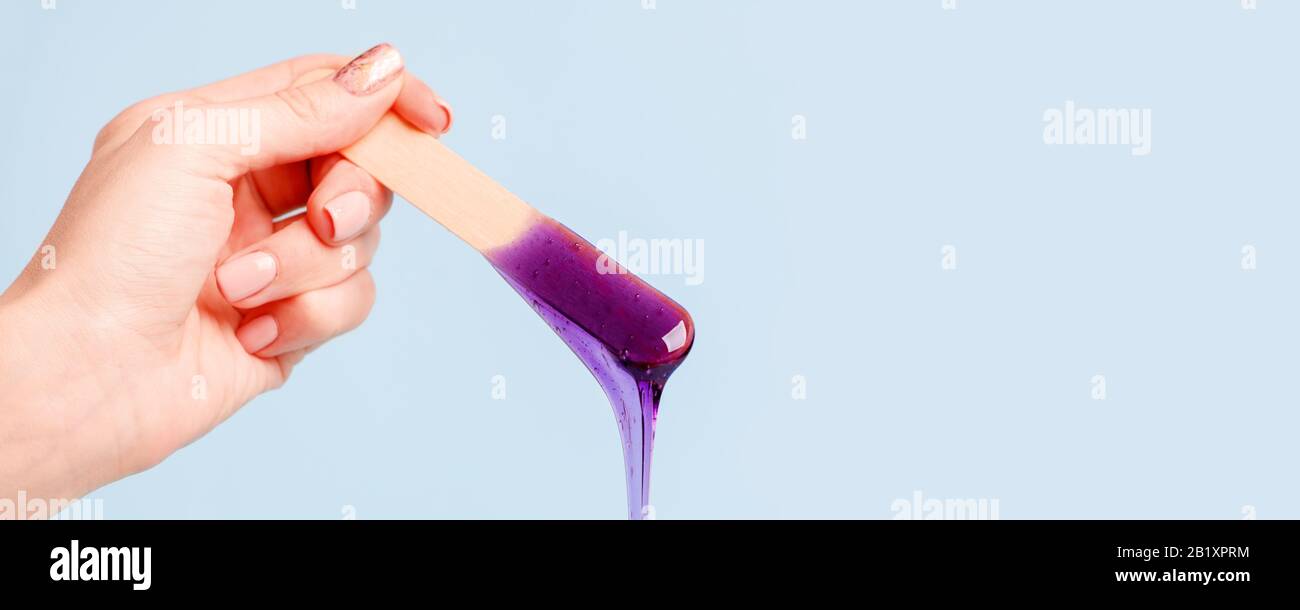 Main tenant la cire violette de beauté ou la pâte de sucre sur la spatule en bois qui coule dans le récipient sur fond bleu. Concept de l'industrie de la beauté publicitaire, l Banque D'Images