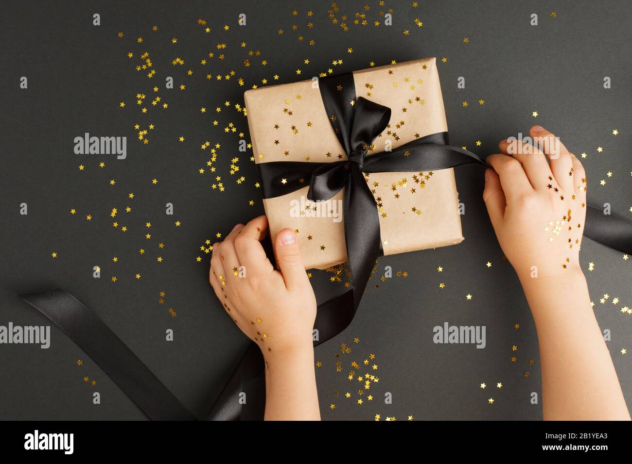 Les mains des enfants tenant la boîte cadeau d'artisanat avec ruban noir sur fond noir avec des confettis d'étoile d'or étincelants. Noël, nouvel an, vente de vendredi noir et Banque D'Images