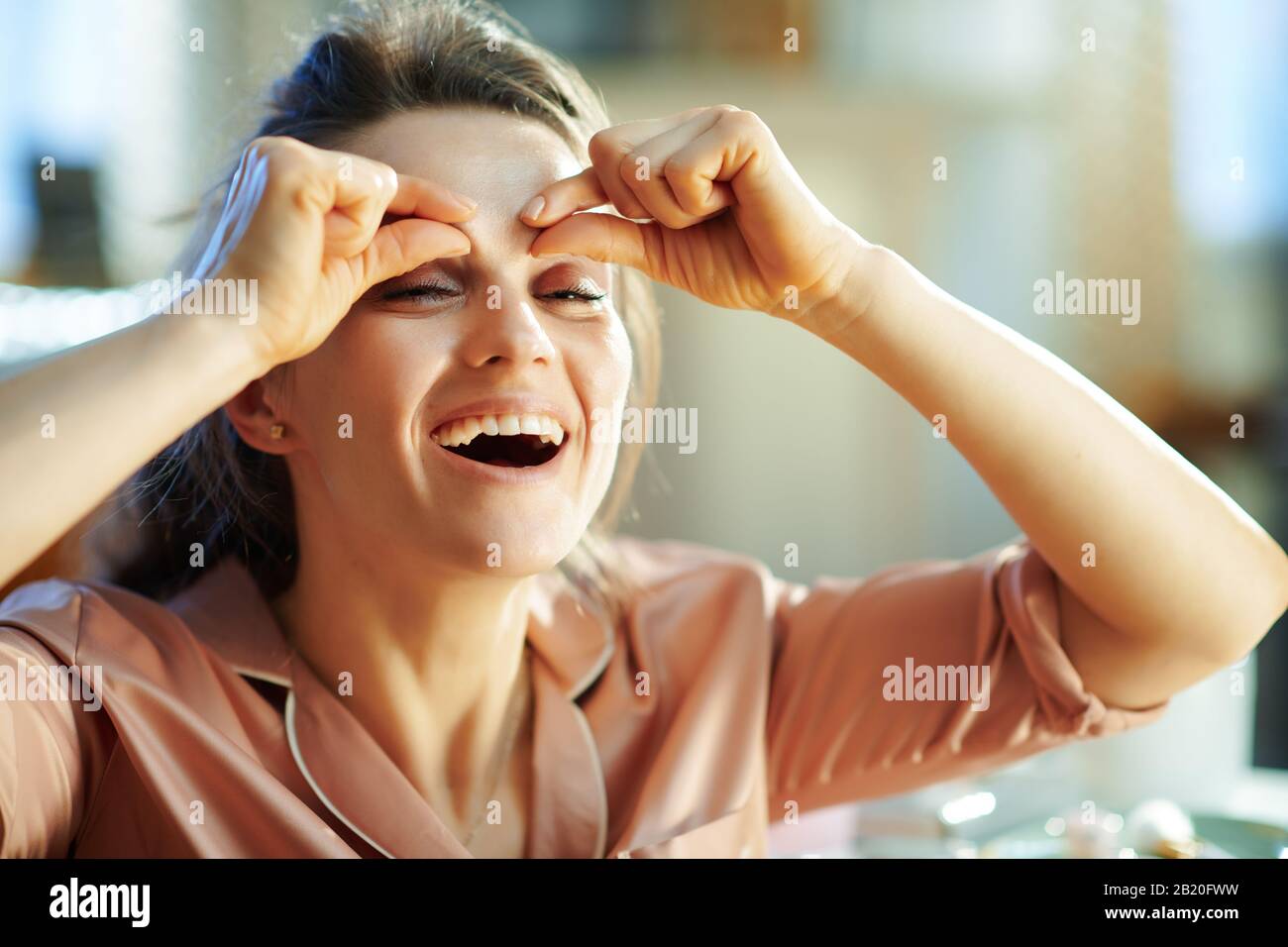 femme de ménage souriante et élégante de 40 ans en pyjama dans le salon moderne en hiver ensoleillé, massage facial. Banque D'Images