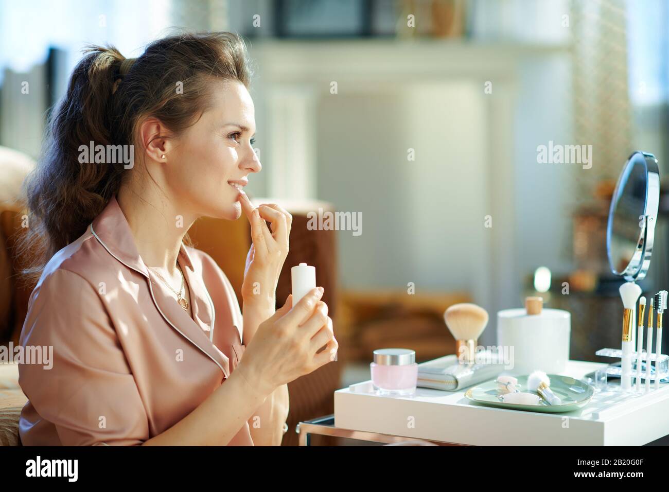 femme de ménage souriante et élégante de 40 ans en pyjama avec tube  cosmétique blanc appliqué à la crème contour des lèvres près de la table  avec des articles de toilette dans