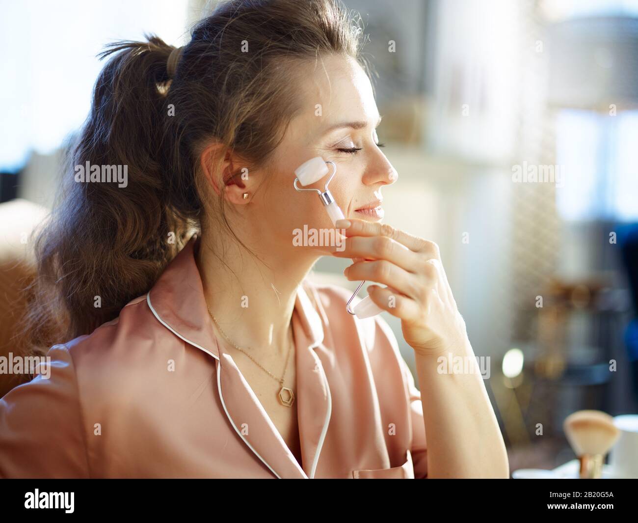 jeune femme détendue en pyjama à la maison moderne en hiver ensoleillé avec masseur à roulettes en quartz apaisant. Banque D'Images