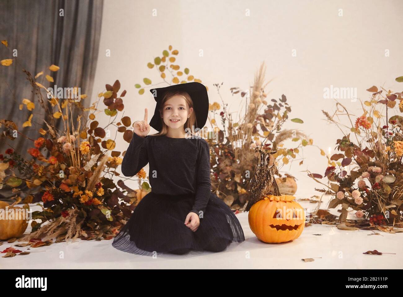 Petite fille mignonne avec de longs cheveux debout sur scène en robe noire et chapeau avec des décorations d'automne sur fond pendant Halloween Banque D'Images