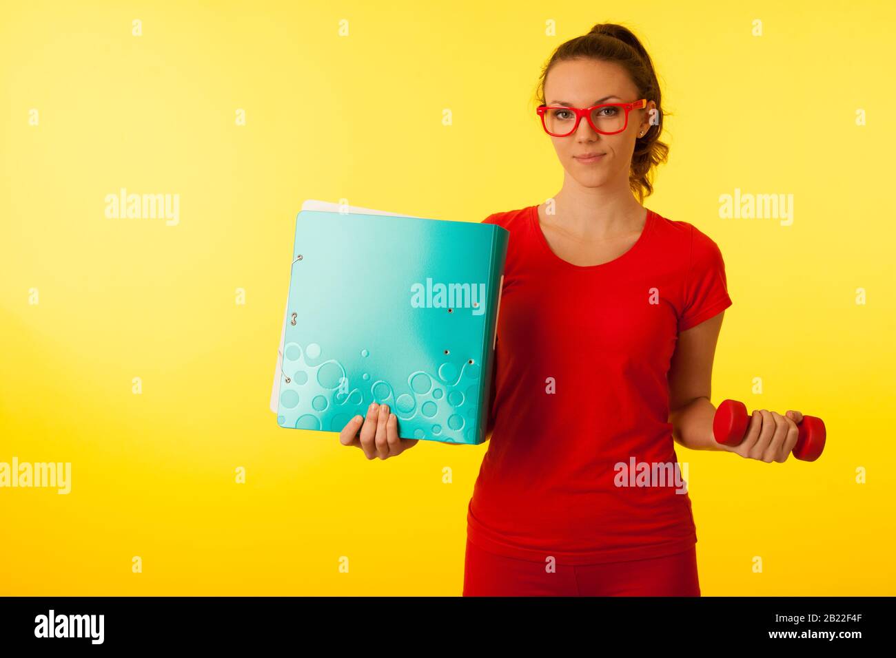 jolie jeune femme caucasienne heureuse dans un t-shirt rouge sur fond jaune vif geste puissance de la connaissance hodling un haltère et un grand dossier Banque D'Images