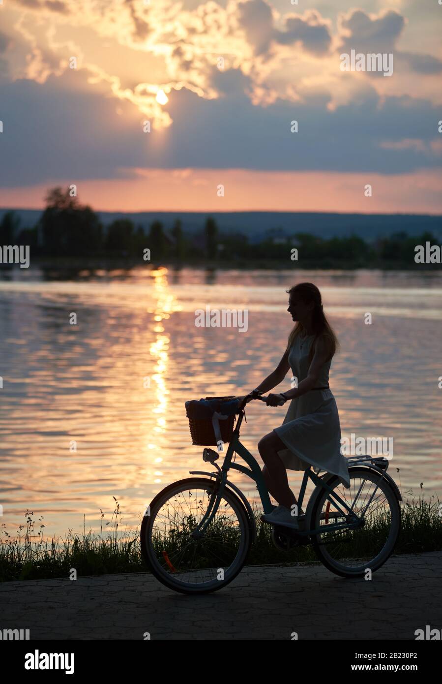 Silhouette d'une jolie fille qui monte un vélo le long du lac au beau coucher du soleil, horizon mauve et chemin du soleil sur l'eau, moments paisibles, espace de copie Banque D'Images