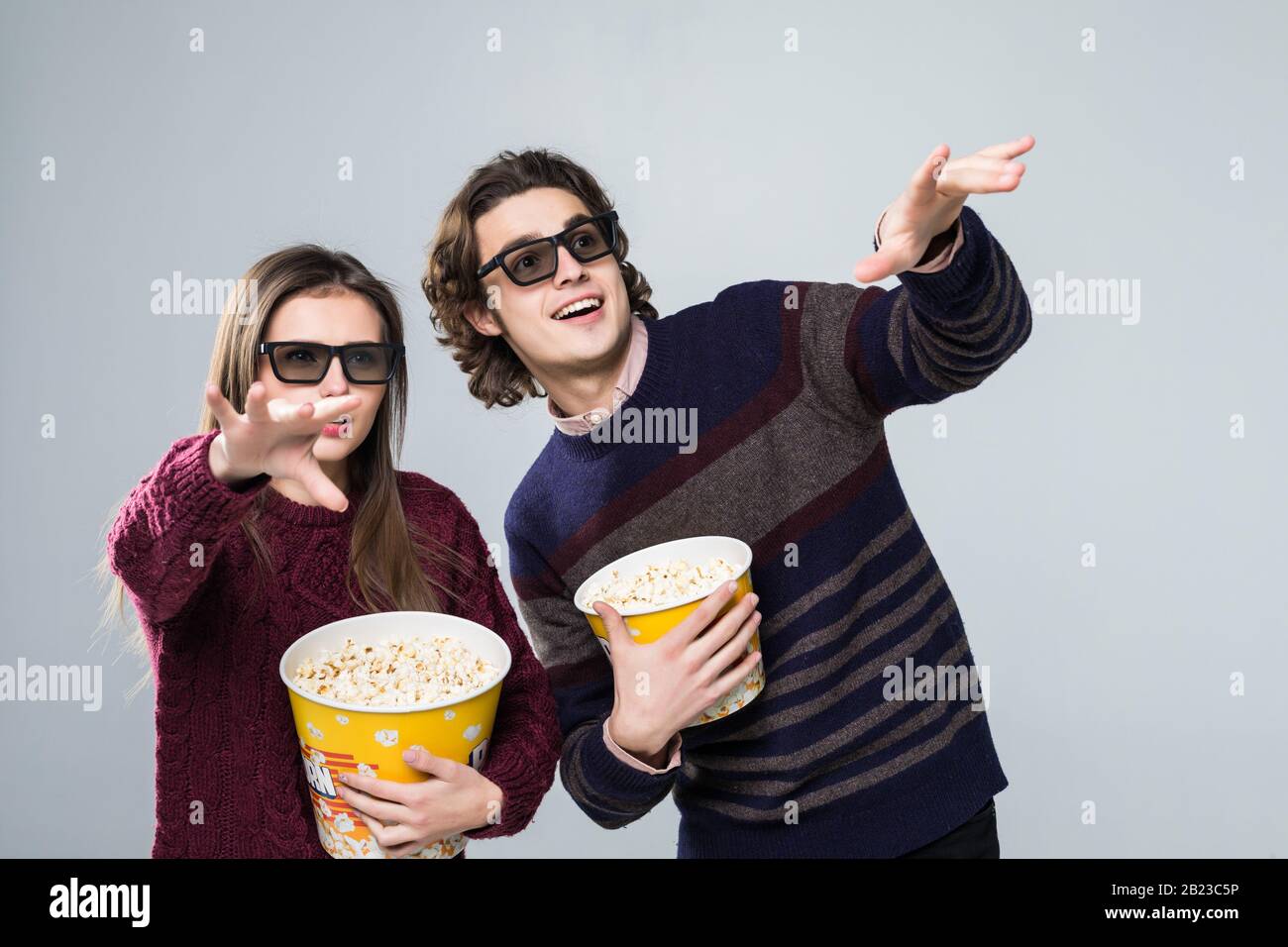 Jeune couple choqué, femme et homme dans des lunettes en relief regardant le film de cinéma à date, tenant seau de popcorn, pointant des index doigts sur le copyspace isolé Banque D'Images
