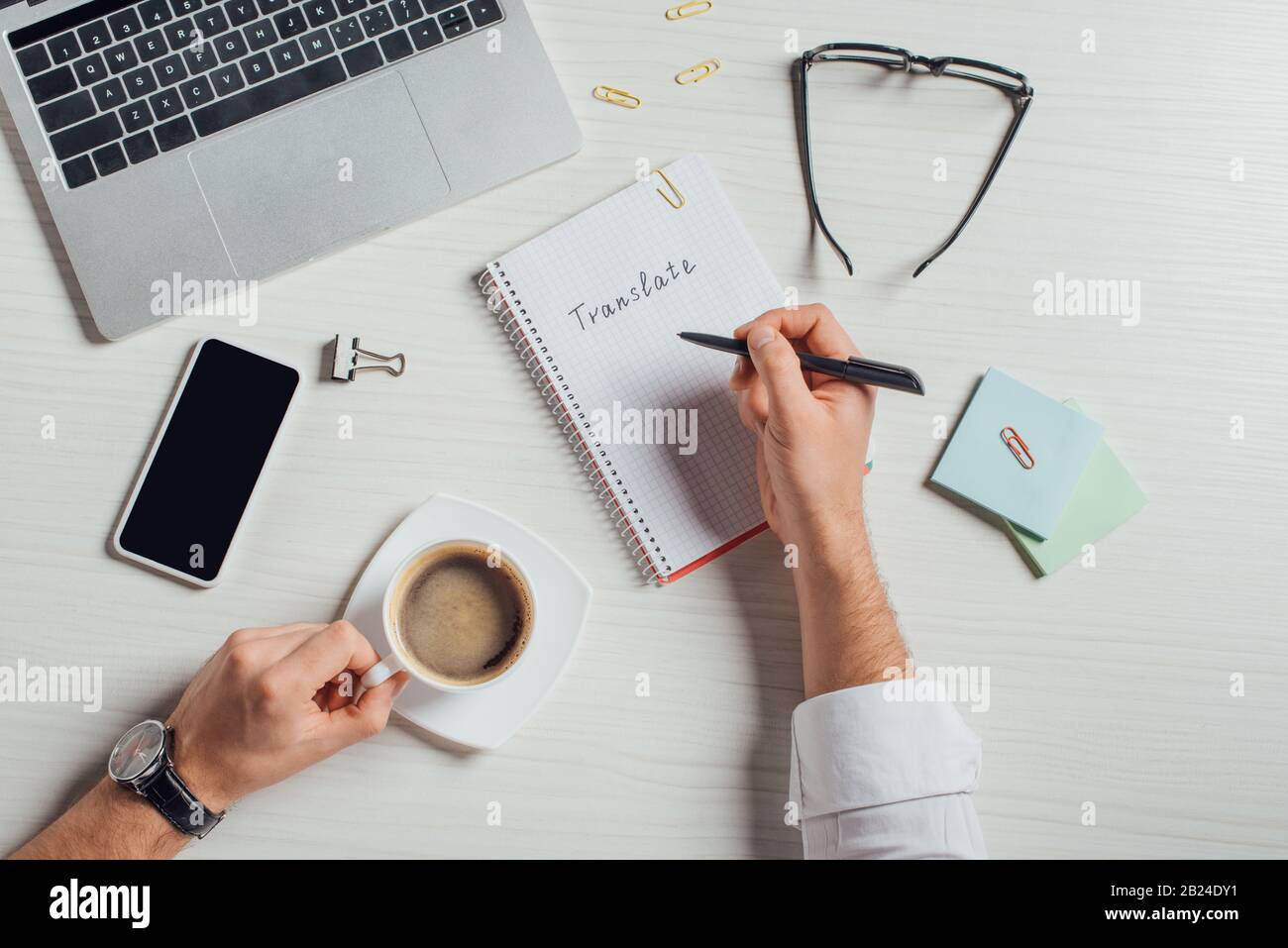 vue rognée du traducteur masculin travaillant avec un ordinateur portable et un smartphone tout en écrivant dans le bloc-notes sur le lieu de travail avec une tasse de café Banque D'Images