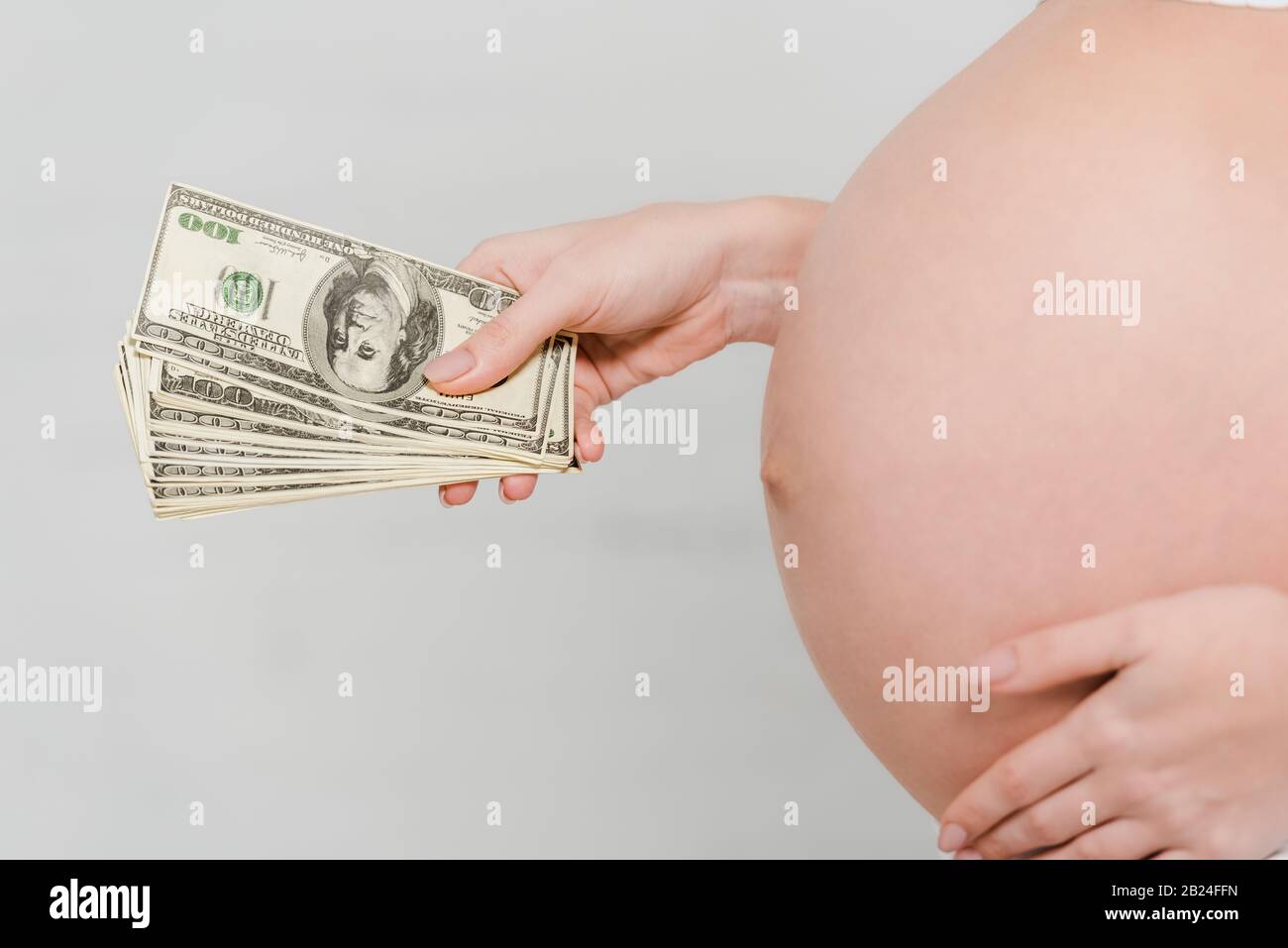 Vue rognée de la femme enceinte qui détient des billets en dollars sur fond gris, concept de maternité de mères porteuses Banque D'Images