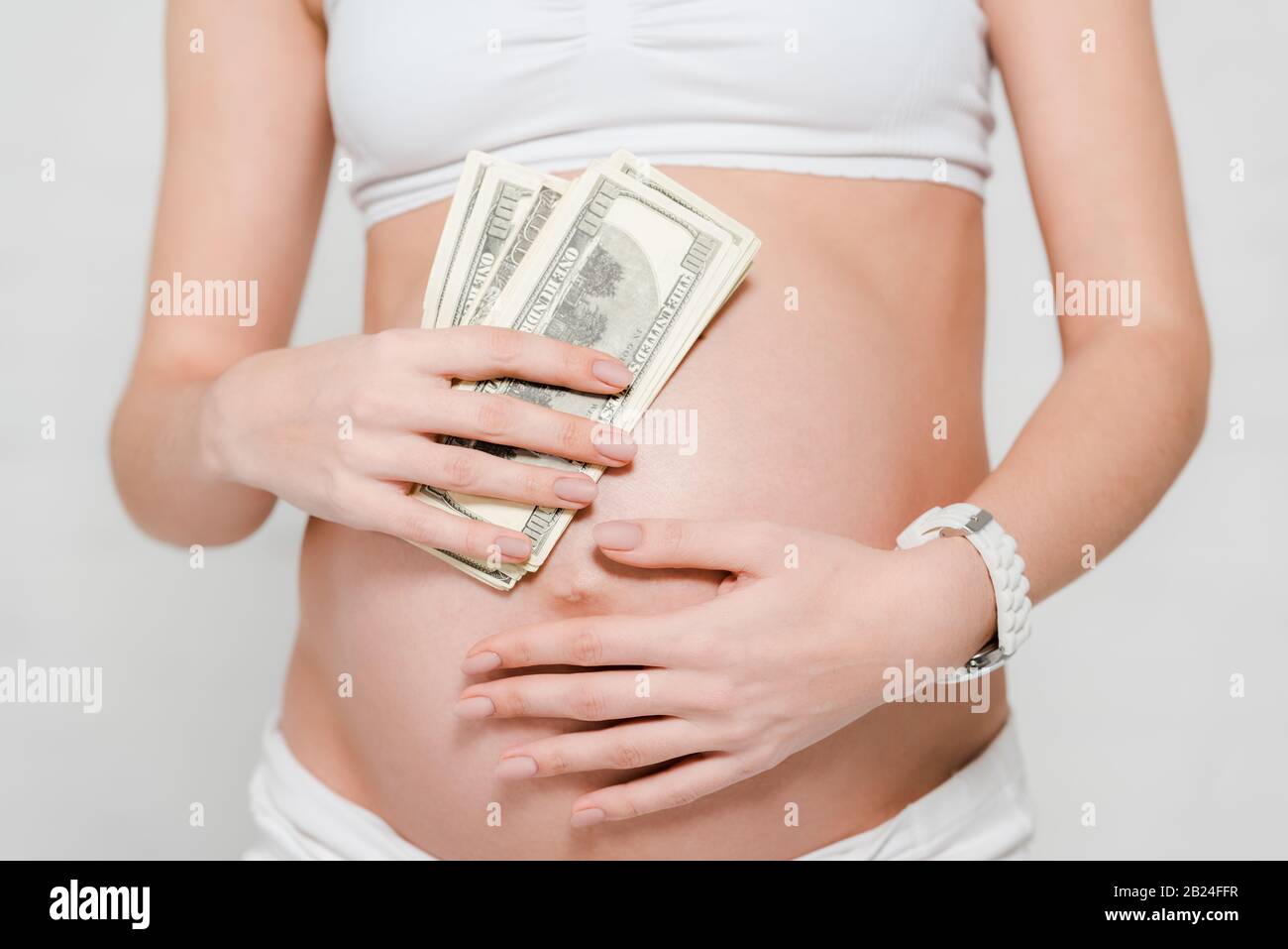 Vue rognée de la femme enceinte qui détient des billets en dollars près du ventre sur fond gris, concept de maternité de mères porteuses Banque D'Images