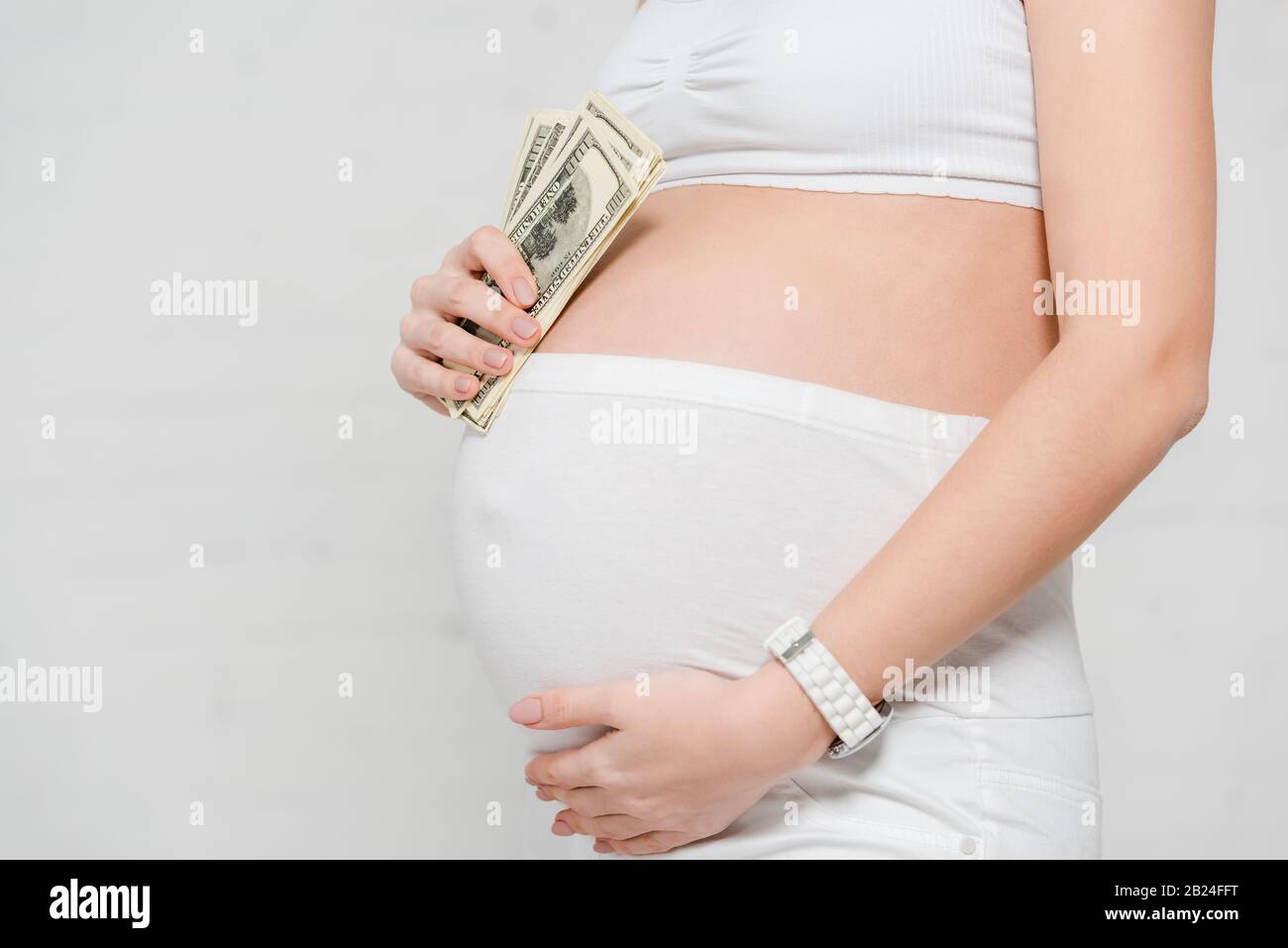 Vue réduite de la femme enceinte qui détient des billets en dollars et qui touche le ventre sur fond gris, concept de maternité de mères porteuses Banque D'Images