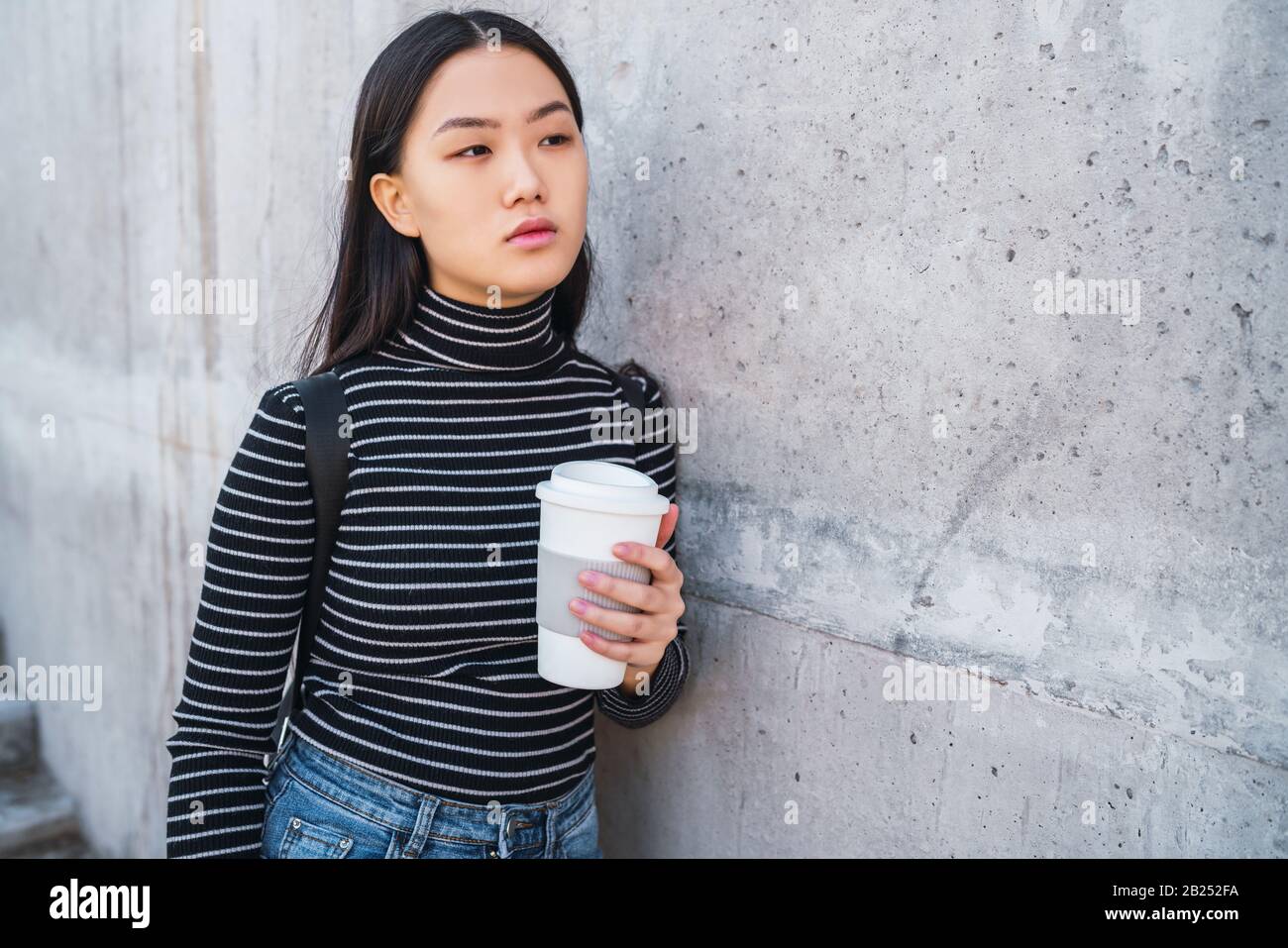 Portrait of young Asian woman holding une tasse de café à emporter contre en béton. Banque D'Images