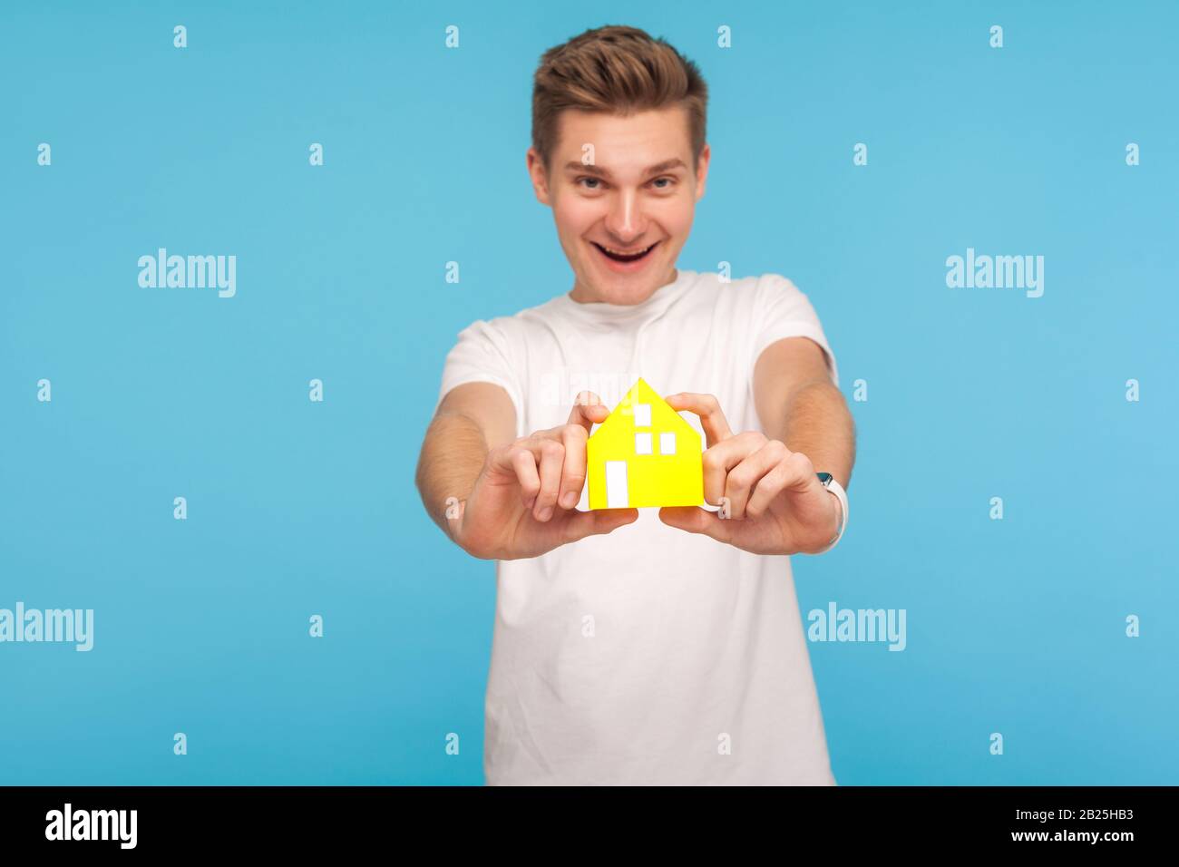 Achat à domicile. Homme joyeux dans un t-shirt blanc tenant une petite maison en papier jaune et souriant à l'appareil photo, assurance immobilière, hypothèque pour les jeunes investisseurs Banque D'Images Achat à domicile. Homme joyeux dans un t-shirt blanc tenant une petite maison en papier jaune et souriant à l'appareil photo, assurance immobilière, hypothèque pour les jeunes investisseurs Banque D'Images