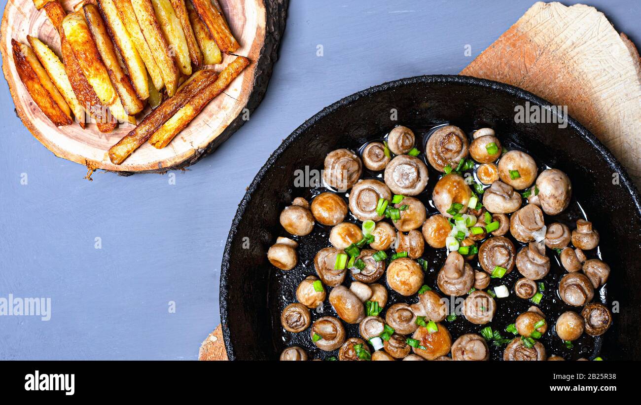 champignons chamignons frits dans une poêle en fonte avec oignons verts et frites sur un support en bois sur fond gris vue de dessus. Banque D'Images