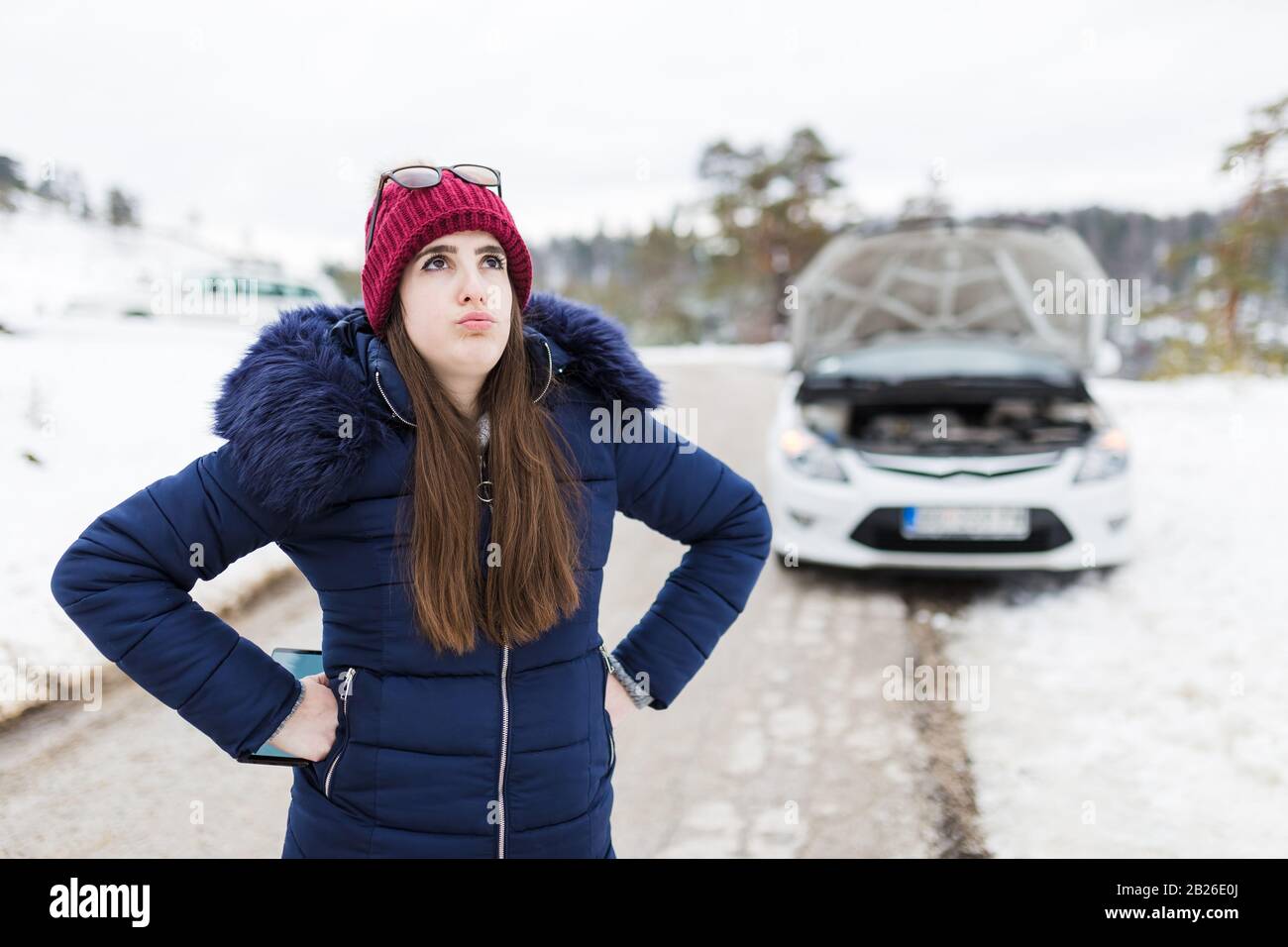 Femme stressée et voiture cassée avec capot ouvert en arrière-plan. Problèmes de déplacement sur route et concepts d'assistance. Banque D'Images